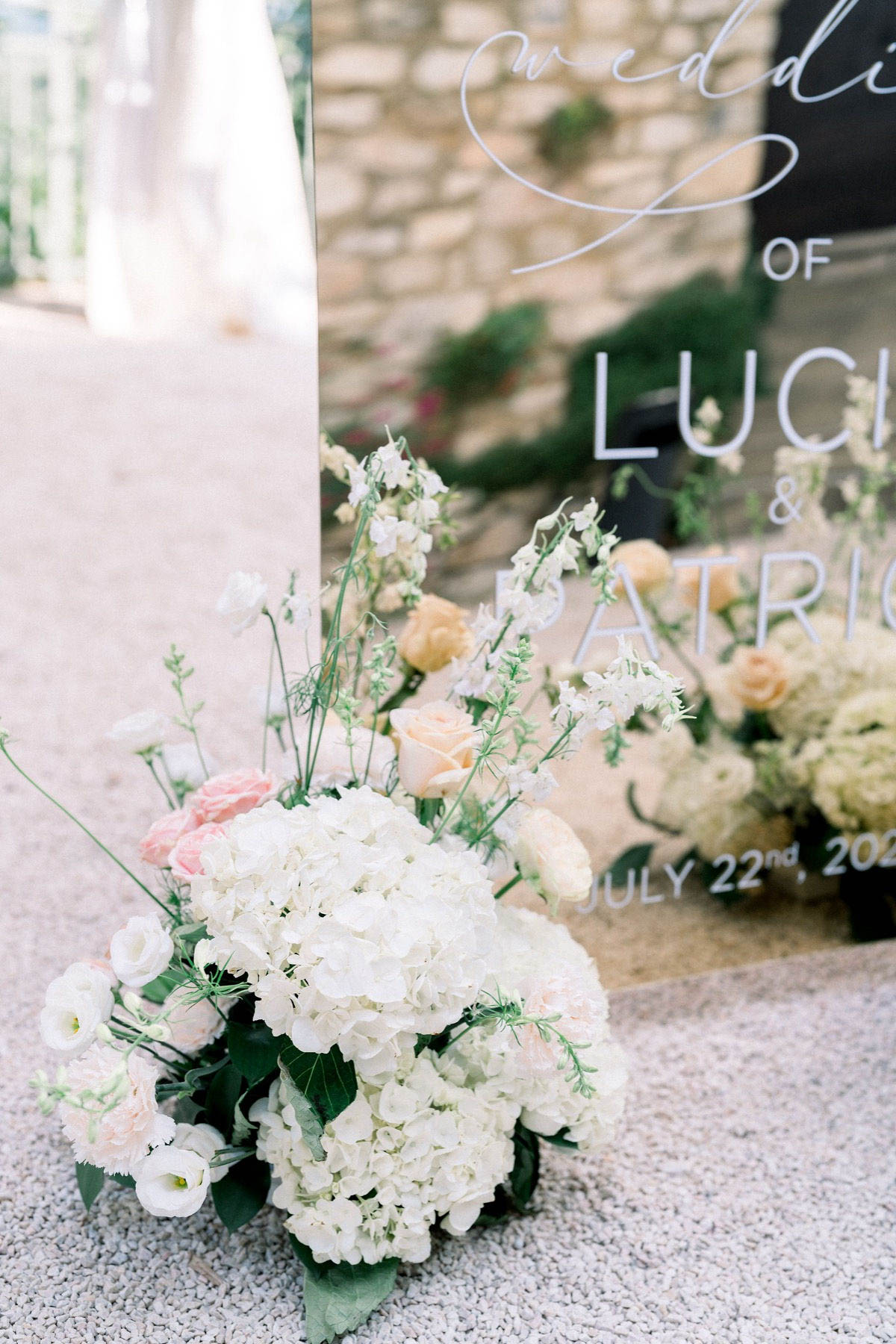 Close-up detail shot of a floral arrangement placed at the base of a mirrored acrylic wedding welcome sign outdoors on a gravel surface, with a stone wall visible in the background. The sign features white hand-lettered calligraphy and block script reading 'Wedding of Luci... & Patric...' with the date 'July 22nd, 20...' The floral arrangement includes white hydrangeas, blush pink roses, peach garden roses, white lisianthus, white stock flowers, and green foliage. The color palette is soft white, blush pink, and peach, consistent with a classic romantic styling theme. A figure in a white dress is partially visible and out of focus at the far left edge of the frame.