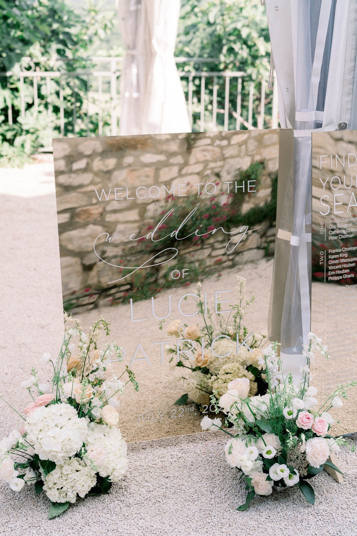Detail shot of a wedding welcome sign displayed outdoors on a gravel surface, with a stone wall and greenery visible in the background. The sign is a large acrylic or mirror panel with white hand-lettered and printed text reading 'Welcome to the Wedding of Lucie & Patrick, July 22nd, 20...' and is partially backed by a sheer grey drape. A seating chart on a matching acrylic panel is partially visible to the right. Four low floral arrangements are placed at the base of the sign, featuring white hydrangeas, blush and cream garden roses, white lisianthus, and delicate green foliage, consistent with a soft, classic palette. The overall styling is modern-classic with a refined, airy aesthetic.