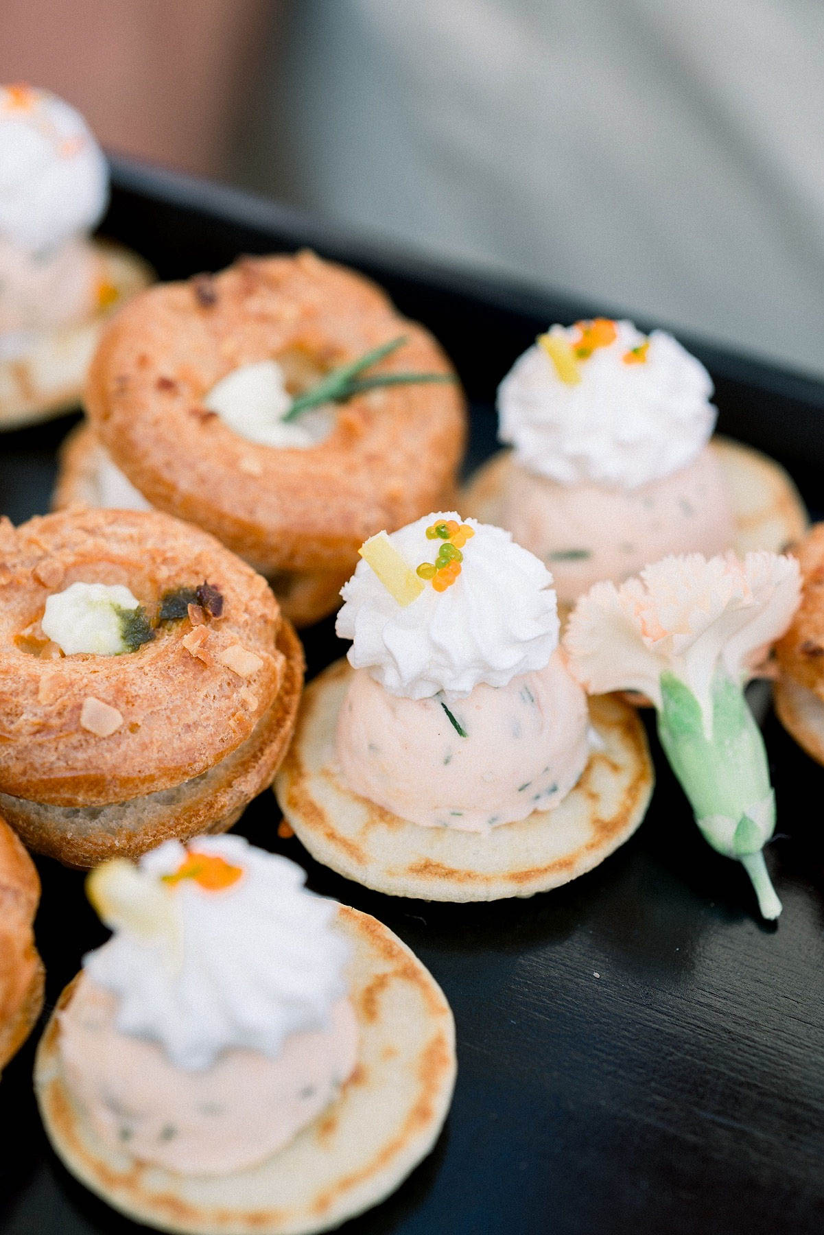 Close-up detail shot of cocktail hour canapés arranged on a black tray. The spread includes two varieties: small blini topped with a pale salmon-pink mousse, a rosette of whipped cream, orange roe, and a sliver of what appears to be candied lemon zest, garnished with a pale peach carnation; and golden-baked choux rings filled with a cream and herb mixture. The presentation reflects a refined catering style with attention to garnish detail, including fresh herb sprigs and orange caviar pearls.