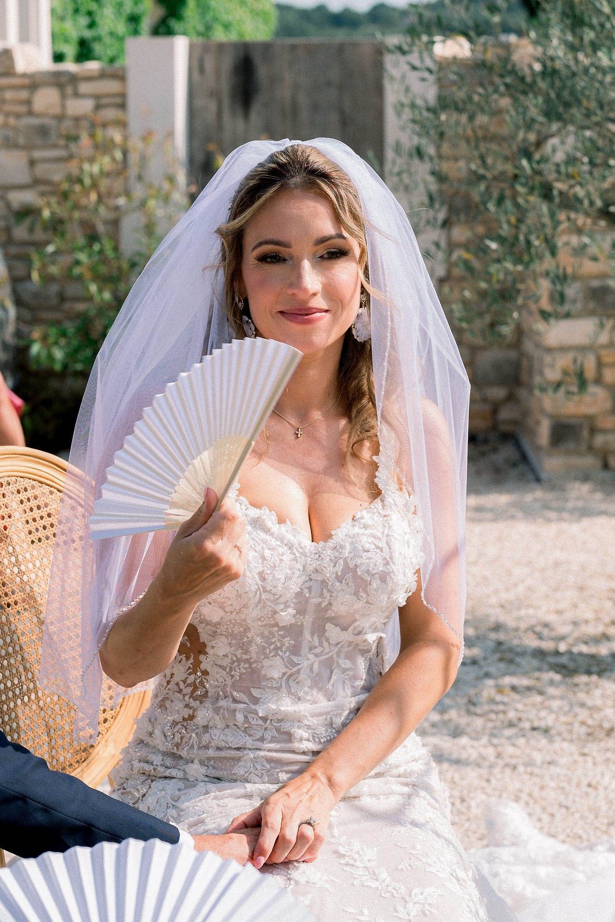 A close-up portrait of a bride seated outdoors during what appears to be a civil or religious ceremony, holding an open ivory paper hand fan, likely to cool herself in the heat. She is wearing a fitted ivory lace gown with a sweetheart neckline featuring floral appliqué detailing, a cathedral-length white tulle veil, chandelier earrings, and a delicate cross necklace. Her hair is styled in loose curls swept to one side, and she wears full glam makeup with defined brows and a warm lip. She is seated on a rattan chair, and a second hand fan is partially visible at the bottom of the frame, suggesting guests were also given fans. The outdoor setting features a gravel courtyard with a stone wall backdrop, consistent with a Provençal or South of France venue.