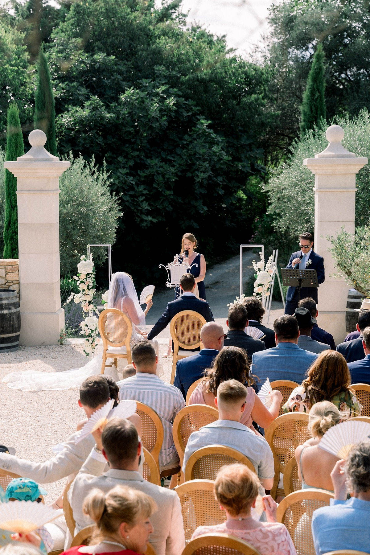 An outdoor wedding ceremony taking place in a formal garden setting with a gravel aisle, framed by two tall stone pillar gate posts. The bride, wearing a white gown and long veil, and the groom in a dark navy suit are seated at the front facing a female officiant in a navy sleeveless dress who is speaking into a microphone at an ornate white lectern; a second person in a navy suit stands at a music stand to the right. Ceremony decor includes tall white floral arrangements on acrylic or glass stands flanking the couple, featuring white blooms and greenery, with additional white floral clusters on an easel-style stand. Approximately 30–40 guests are seated in rows of natural rattan cane-back chairs facing the ceremony, and many guests are holding white handheld paper fans, suggesting warm weather. The wide-angle shot is taken from behind the seated guests looking toward the altar area, giving a full view of the ceremony layout and garden backdrop.