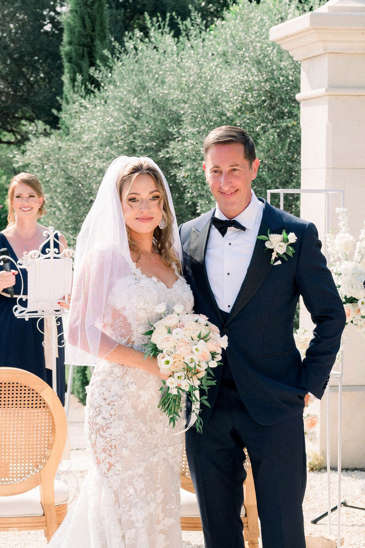The bride and groom pose together immediately following an outdoor ceremony, likely at a French chateau or estate, with a limestone pillar and olive trees visible in the background. The bride wears a fitted, heavily embellished lace gown with a mid-length blush-tinted veil and holds a lush bouquet of blush garden roses, white ranunculus, and trailing greenery. The groom is dressed in a navy tuxedo with black bow tie and a white floral boutonniere featuring small white blooms and greenery. In the background, a woman in a navy dress stands near an ornate white iron music stand, and an acrylic ceremony structure with white floral arrangements is partially visible to the right; cane-back chairs used for guest seating are also visible. The shot is a medium portrait framing both subjects from approximately the waist up, with a classic, formal styling aesthetic.
