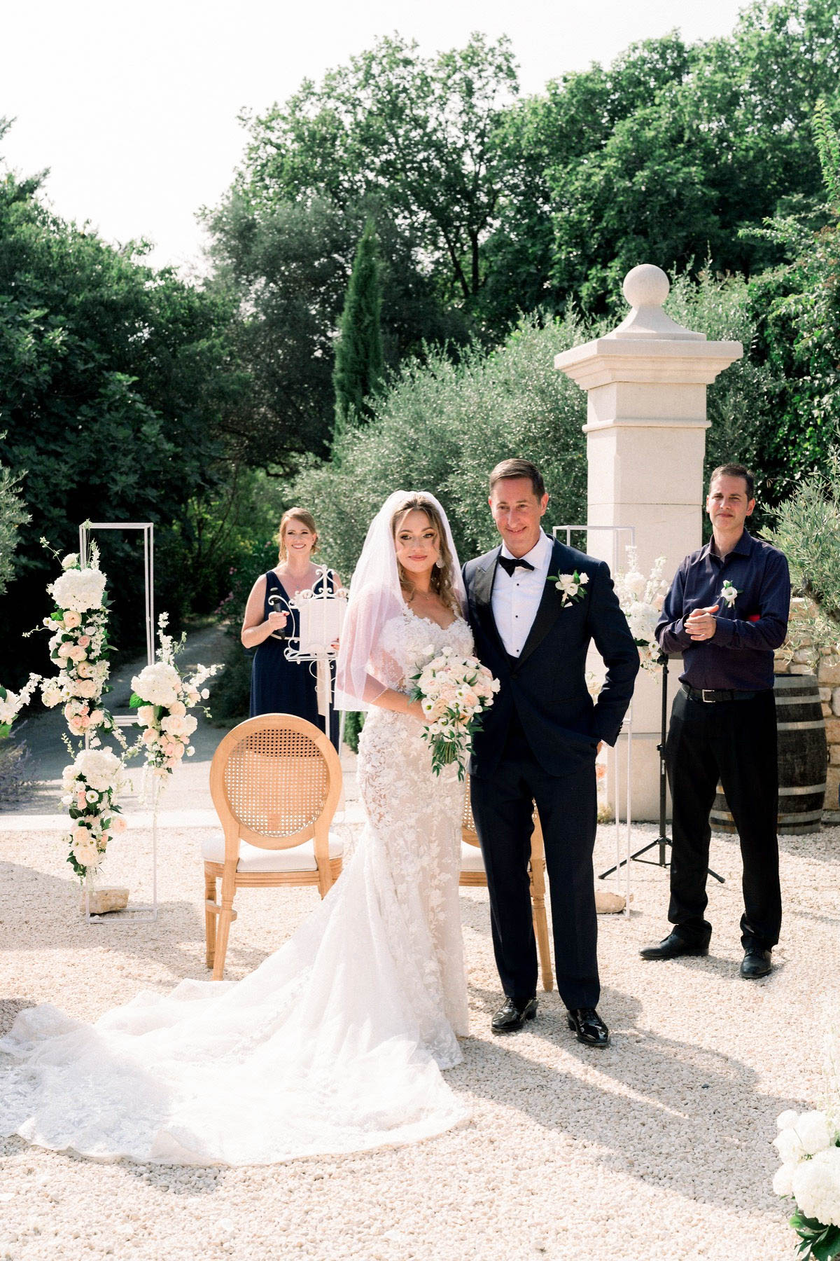 An outdoor wedding ceremony moment captured at what appears to be a French chateau or domaine, with the bride and groom standing together on a gravel courtyard beside a classical stone pillar gate. The bride wears a fitted lace mermaid-style gown with a long cathedral train and a blush-toned veil, holding a bouquet of ivory and blush blooms including hydrangeas and roses with greenery. The groom wears a dark navy tuxedo with a black bow tie and a white boutonniere. Behind them, a female officiant in a navy dress holds a ceremony book at a decorative wrought-iron stand, while a male sound technician stands to the right near audio equipment. The ceremony setup includes acrylic stand-alone frames flanked by tall floral arrangements of white hydrangeas, blush roses, and trailing greenery, alongside a cane-back Louis XVI-style chair. The overall styling is classic and refined with a blush and ivory floral palette. Medium wide portrait shot.