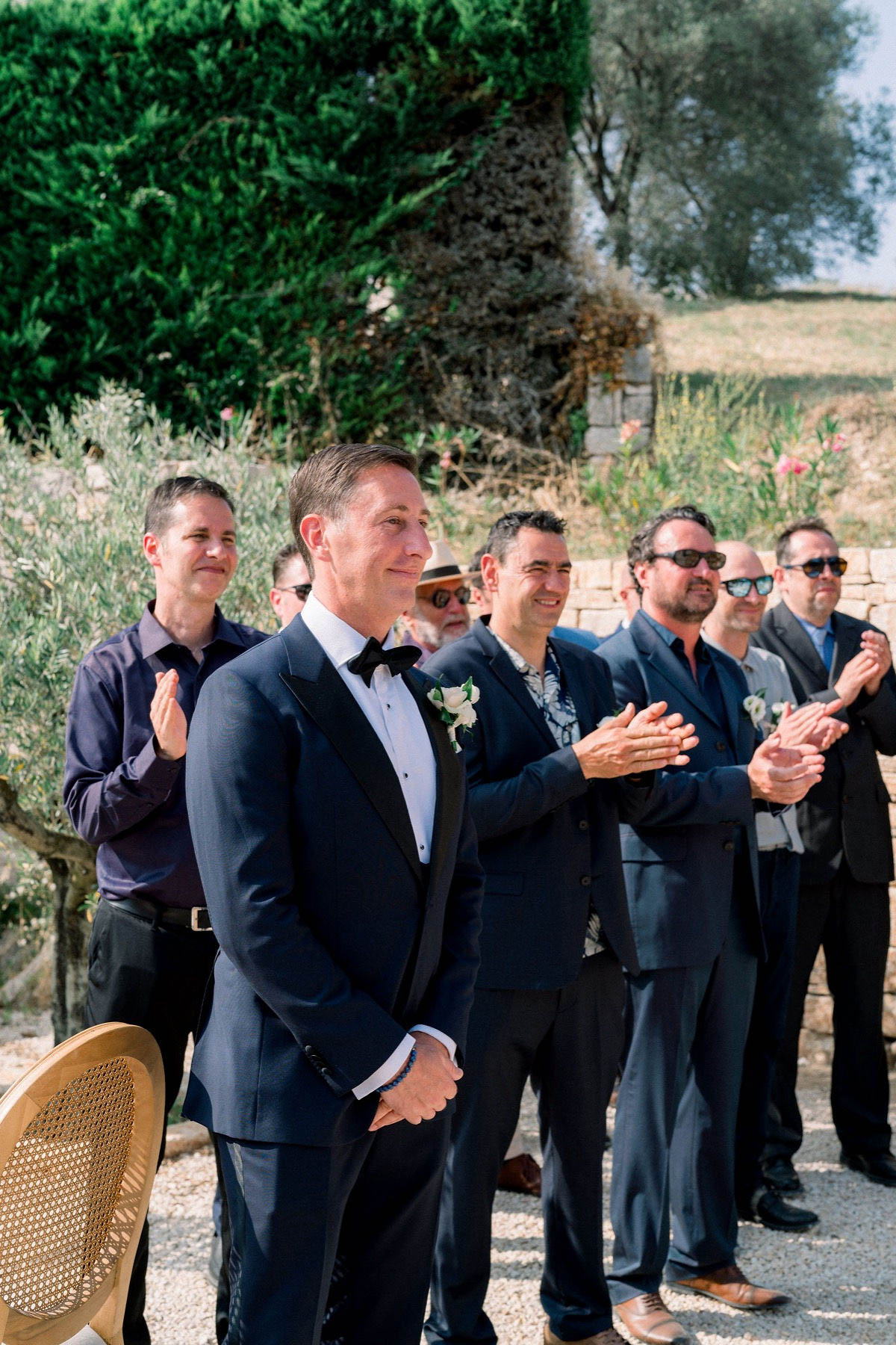 An outdoor daytime wedding ceremony showing the groom standing at the altar watching the bride's processional, with approximately six male guests standing and applauding behind him. The groom wears a navy tuxedo with black satin lapels, a black bow tie, and a white rose boutonniere with greenery. The guests are dressed in a mix of navy suits, a gray suit, and smart casual attire. The ceremony takes place on a gravel surface with rattan gold chairs visible, set against a low stone wall and Mediterranean-style landscaping. The shot is a portrait-oriented medium shot focused on the groom's emotional expression as he looks toward the approaching bride.