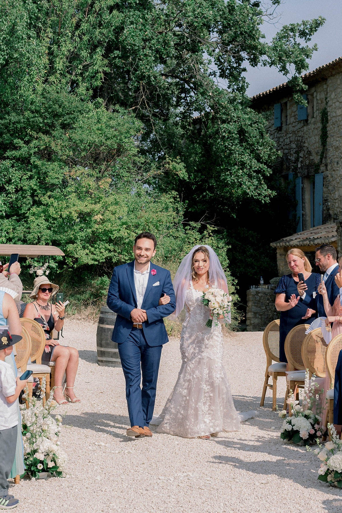 The bride and groom walk back down the aisle together following an outdoor wedding ceremony at what appears to be a French stone farmhouse or mas, identifiable by its light-colored stone facade and blue shutters visible in the background. The groom wears a navy blue suit with a fuchsia boutonniere and tan loafers, while the bride wears a fitted lace gown with floral appliqué detailing, a cathedral-length veil, and carries a bouquet of ivory and white blooms including what appear to be roses and ranunculus. Guests seated in gold cane-back chairs line the gravel aisle on both sides, many holding up phones to photograph the couple, with white and green floral arrangements marking the aisle ends. The overall decor palette is white, ivory, and green with a classic French countryside aesthetic, captured in a mid-distance portrait shot.