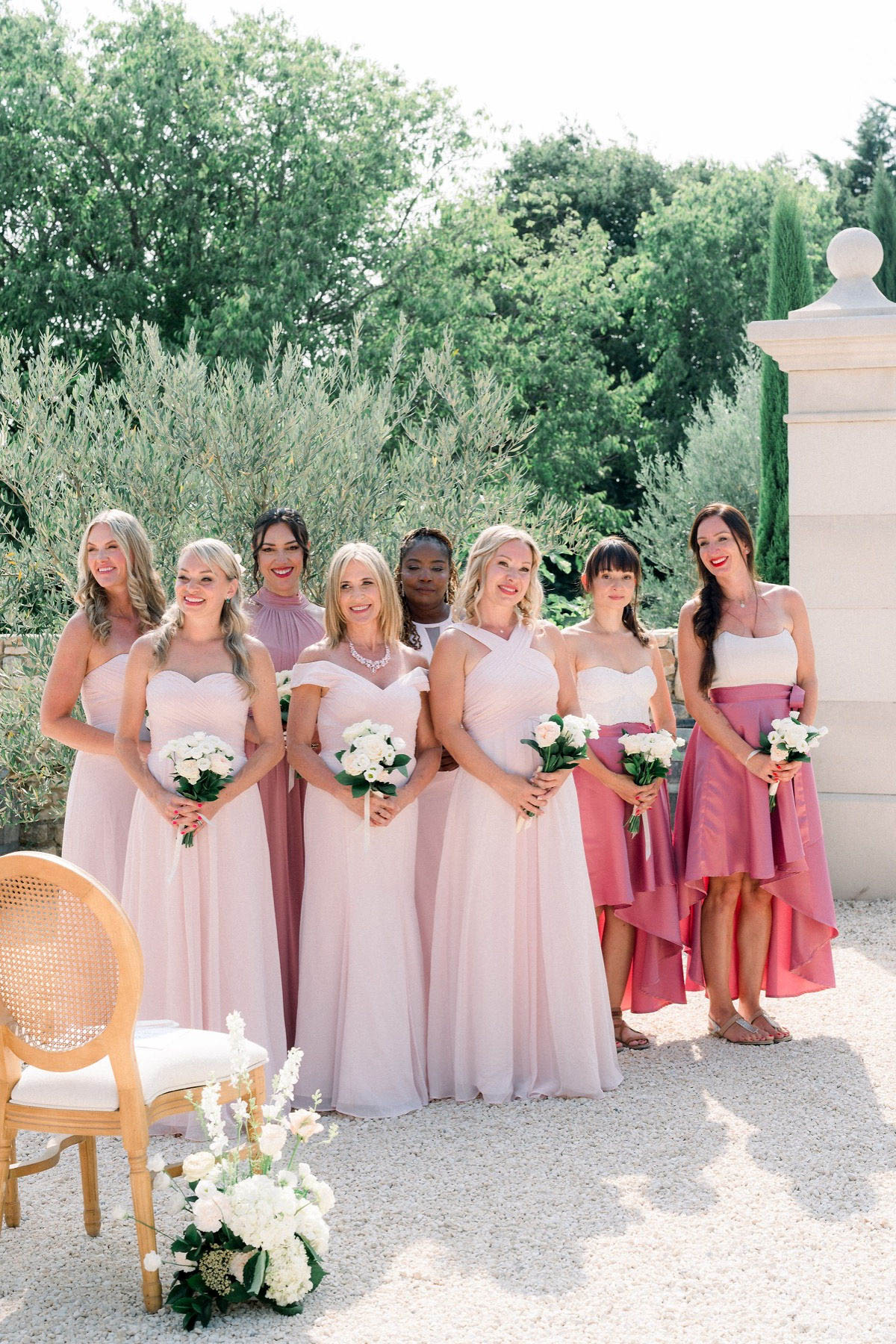 A group portrait of seven bridesmaids standing together outdoors during a wedding ceremony, positioned on a gravel surface beside a stone pillar and a gold cane-back chair. Four of the women wear floor-length blush pink dresses in varying neckline styles — off-the-shoulder, halter, and strapless — while one wears a mauve high-neck dress, one wears a white crop top with a rose-pink hi-low ruffled skirt, and one in the back wears a white top. Each bridesmaid holds a small bouquet of white flowers, and a larger white floral arrangement with dark green foliage sits at ground level near the ceremony chair in the foreground. The styling is classic and cohesive with a pink-toned color palette across mismatched dress styles. The shot is a medium full-length group portrait taken in natural daylight.