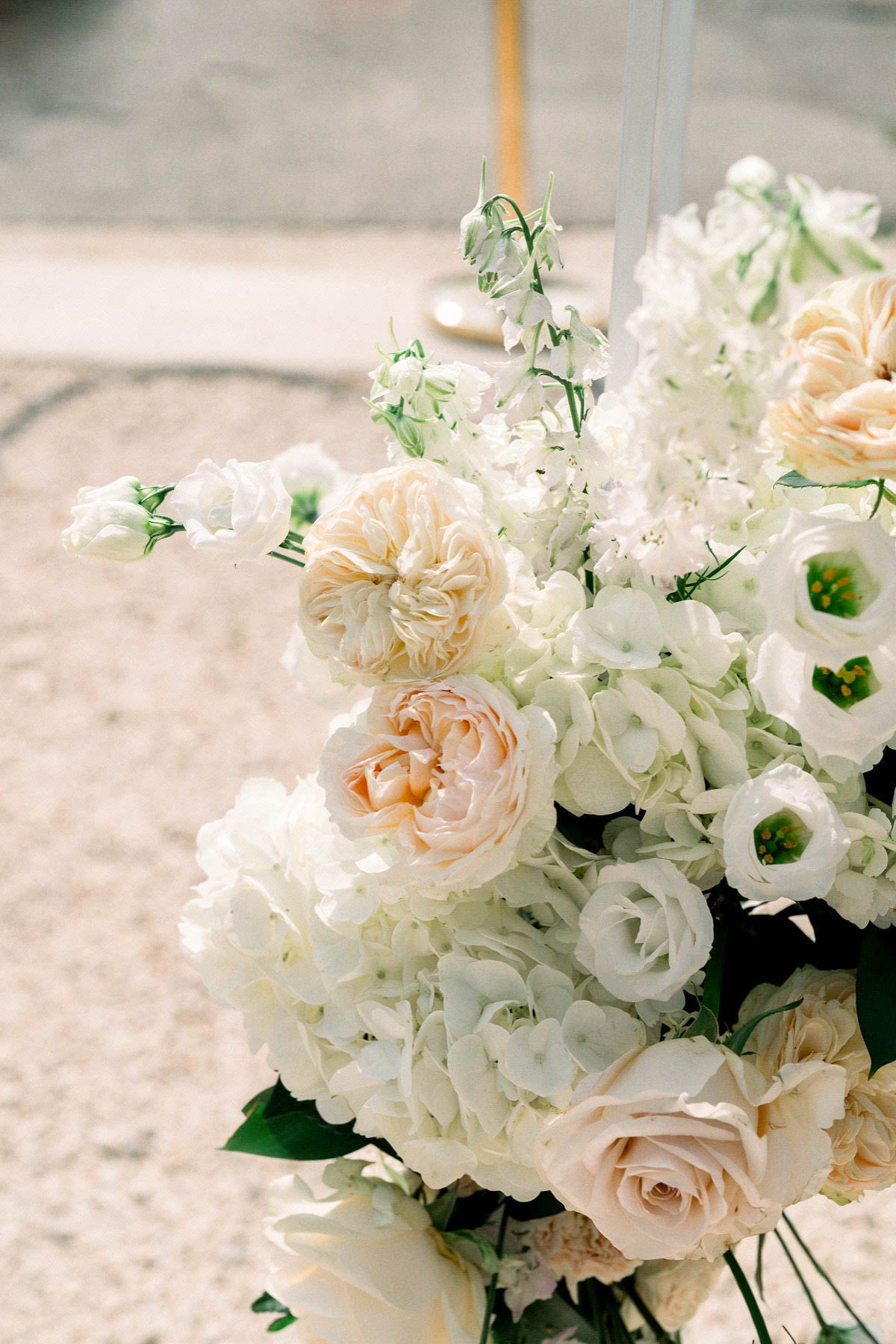 Close-up detail shot of a large floral arrangement, likely positioned at a ceremony or reception setting with a gravel surface visible in the blurred background. The arrangement features white hydrangeas as the base, blush and peach garden roses, white lisianthus with yellow-green centers, white delphinium spires, and blush standard roses, all in a soft white and blush peach color palette with dark green foliage accents. A clear acrylic or glass structural element is partially visible behind the arrangement, suggesting a modern arch or stand. The composition is tightly framed to highlight the floral detail and texture.