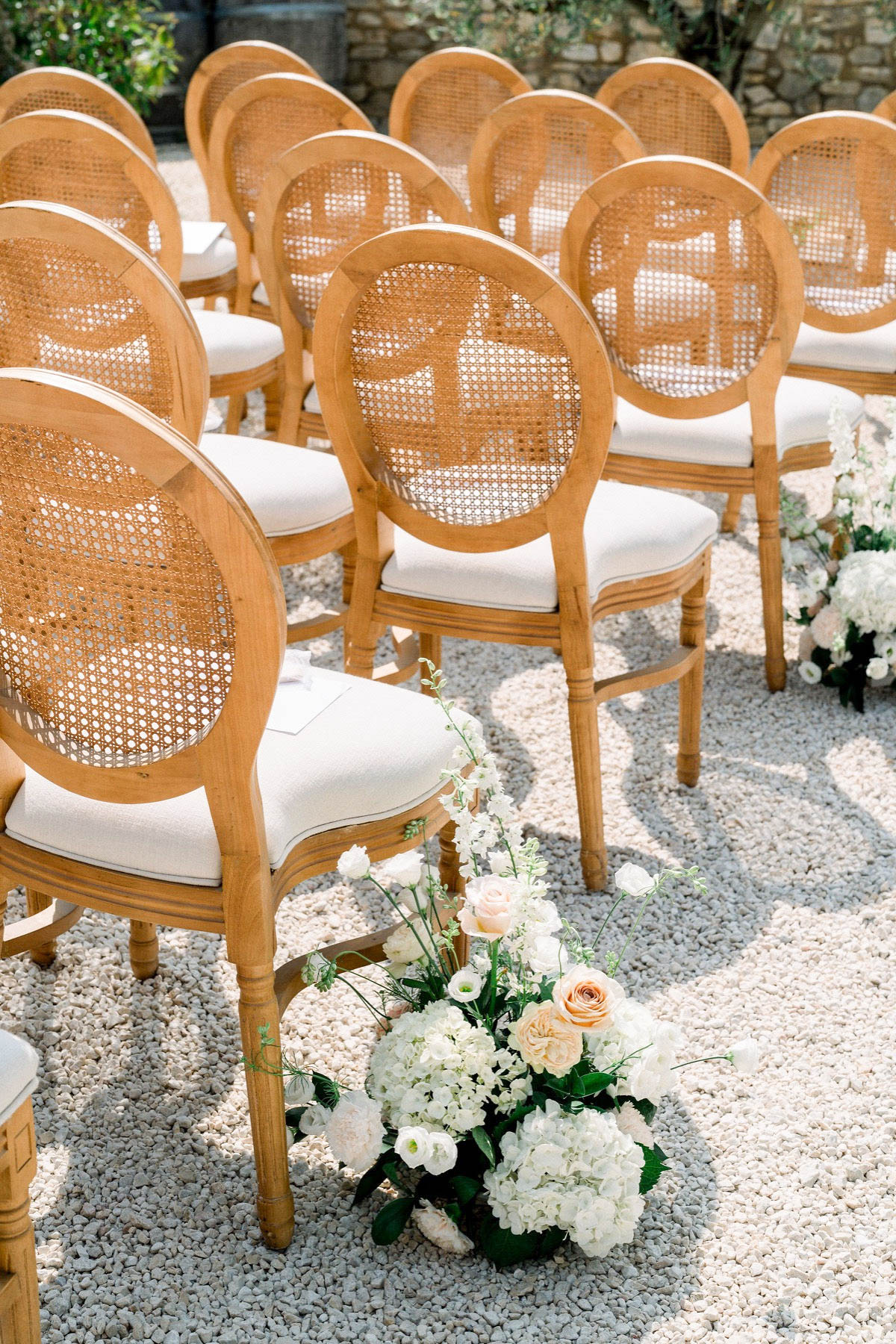 An outdoor ceremony setup shot showing rows of natural wood Louis XVI-style chairs with cane medallion backs and ivory cushioned seats, arranged on a gravel surface. Floral arrangements placed along the aisle feature white hydrangeas, white lisianthus, blush and peach roses, and green foliage. The decor palette is white, blush, and peach with warm natural wood tones, fitting a classic French garden ceremony aesthetic. The composition is a medium angled shot focused on chair and aisle details with no people present.