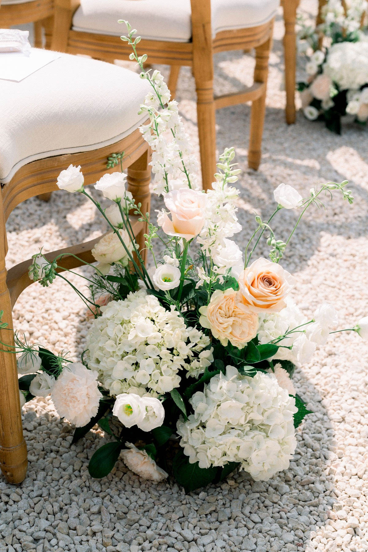 A close-up detail shot of an outdoor ceremony aisle floral arrangement placed on a gravel surface beside natural wood chairs with cream linen cushions. The floor-level arrangement features white hydrangeas as the base, accented with peach garden roses, blush carnations, white lisianthus, white delphinium spires, and dark green foliage. The color palette is white, cream, and soft peach against deep green leaves. Additional floral arrangements are visible in the background along the ceremony aisle, suggesting a classic, romantic ceremony setup.