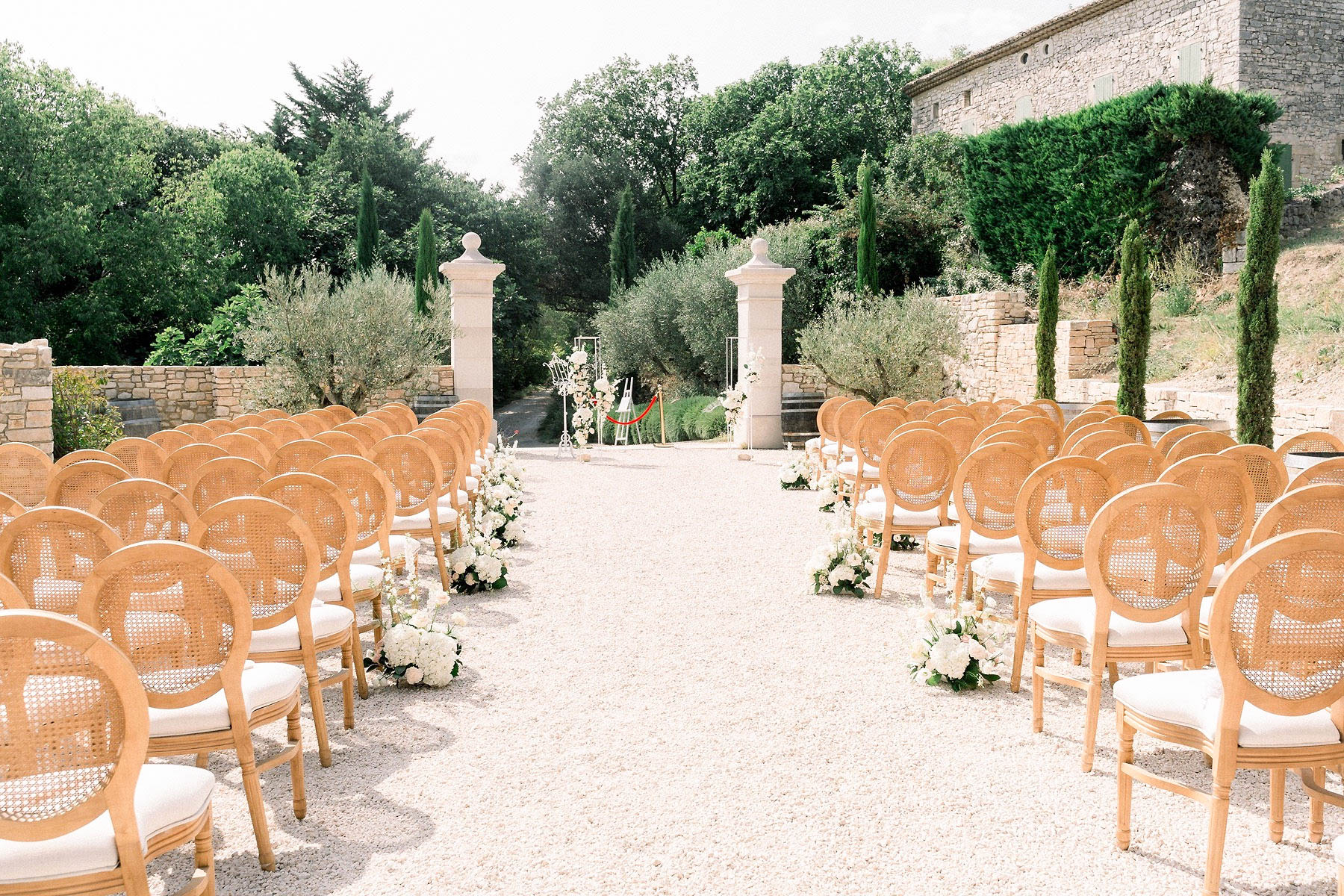 An outdoor wedding ceremony setup on a gravel courtyard at a French stone mas or bastide property, photographed before guests arrive. Rows of natural wood cane-back Louis XVI-style chairs with ivory cushions are arranged on either side of a wide aisle, accommodating approximately 80–100 guests. White floral arrangements of what appear to be white hydrangeas, garden roses, and greenery are placed at the end of several aisle rows as ground-level clusters. At the far end of the aisle, two tall stone pillars frame the ceremony area, with a white arch or altar structure adorned with white florals visible in the background. The overall decor palette is natural wood and ivory with an all-white floral scheme, giving a classic Provençal aesthetic. Wide shot taken from the back of the ceremony space looking toward the altar. Potential venue feature image.