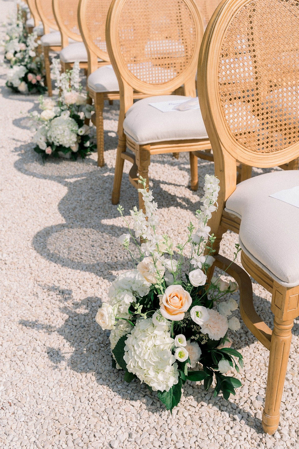 Detail shot of an outdoor wedding ceremony setup on a pale gravel surface, showing a row of natural wood French Louis-style chairs with cane backs and cream upholstered seat cushions. Aisle floral arrangements are placed at the base of each chair, consisting of white hydrangeas, white spray roses, white lisianthus, white delphinium, a peach garden rose, and dark green foliage. The closest arrangement is in sharp focus while those receding down the aisle are softly blurred, creating depth. A printed ceremony program is visible on one of the chair seats. The overall decor palette is cream, white, and soft peach with a classic French-inspired styling.