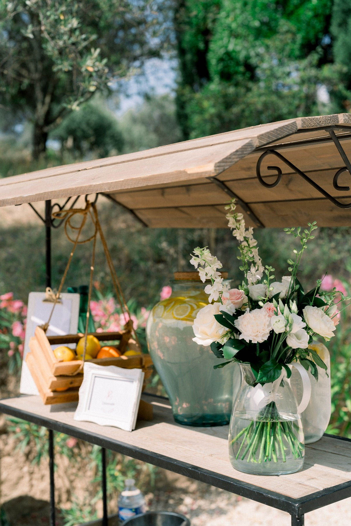 Close-up detail shot of an outdoor cocktail hour or welcome station set up in a garden setting. The display features a rustic wooden and wrought-iron shelving unit with a canopy roof, styled with a clear glass pitcher holding a loose bouquet of cream roses, blush spray roses, white lisianthus, and greenery. A large sage-green ceramic urn with lemon motifs sits beside the bouquet, along with a wooden crate filled with lemons and oranges suspended from the canopy frame, a small white ornate picture frame with a handwritten card, and stacked wooden trays. The overall styling palette is soft and rustic-Mediterranean, combining muted sage, cream, and blush tones with natural wood and ironwork elements.