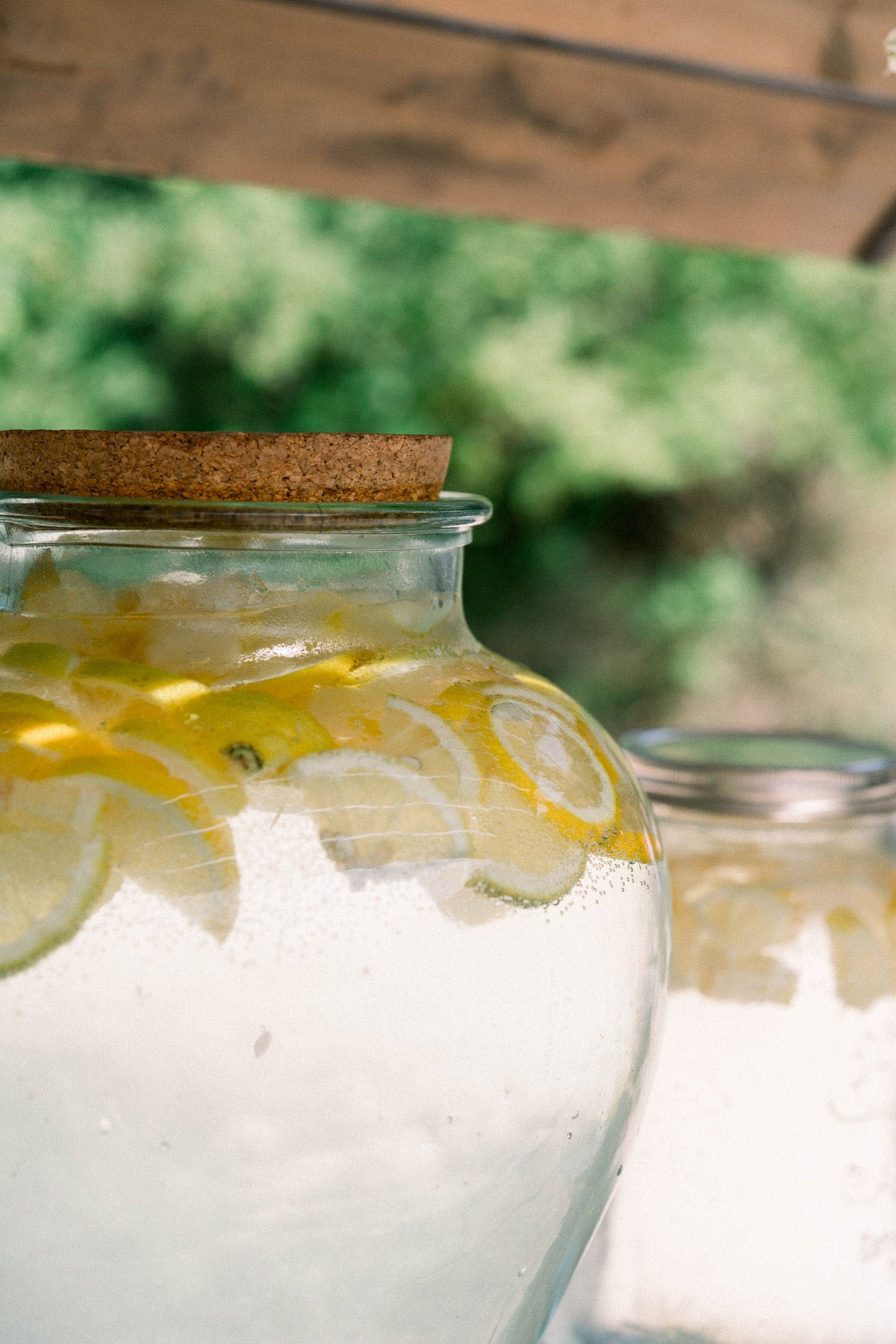Close-up detail shot of a large round glass carafe with a cork stopper, filled with a clear lemonade or infused water featuring visible slices of yellow lemon and lime. A second similar glass jar is partially visible and out of focus in the background to the right. The setting appears to be an outdoor cocktail hour or drinks station, with blurred greenery visible in the background. The styling is casual and rustic, consistent with a relaxed outdoor summer wedding aesthetic.