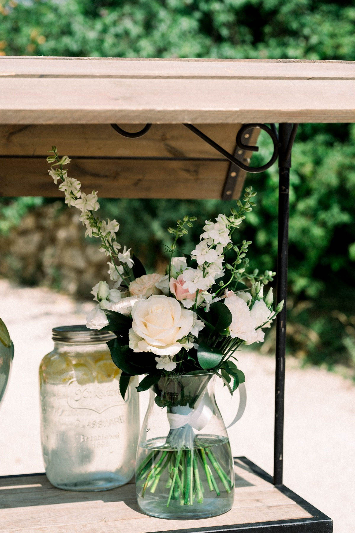 Close-up detail shot of a rustic outdoor wedding decor setup, showing a small floral arrangement in a clear glass pitcher placed on a wooden shelf unit with black iron brackets. The bouquet includes cream roses, blush pink roses, white stock flowers, white lisianthus, and dark green foliage, tied with a white ribbon. Beside it sits a large mason jar filled with water and lemon slices, suggesting a drinks or welcome station. The styling is rustic and relaxed, with natural wood, iron hardware, and simple glassware forming the palette.