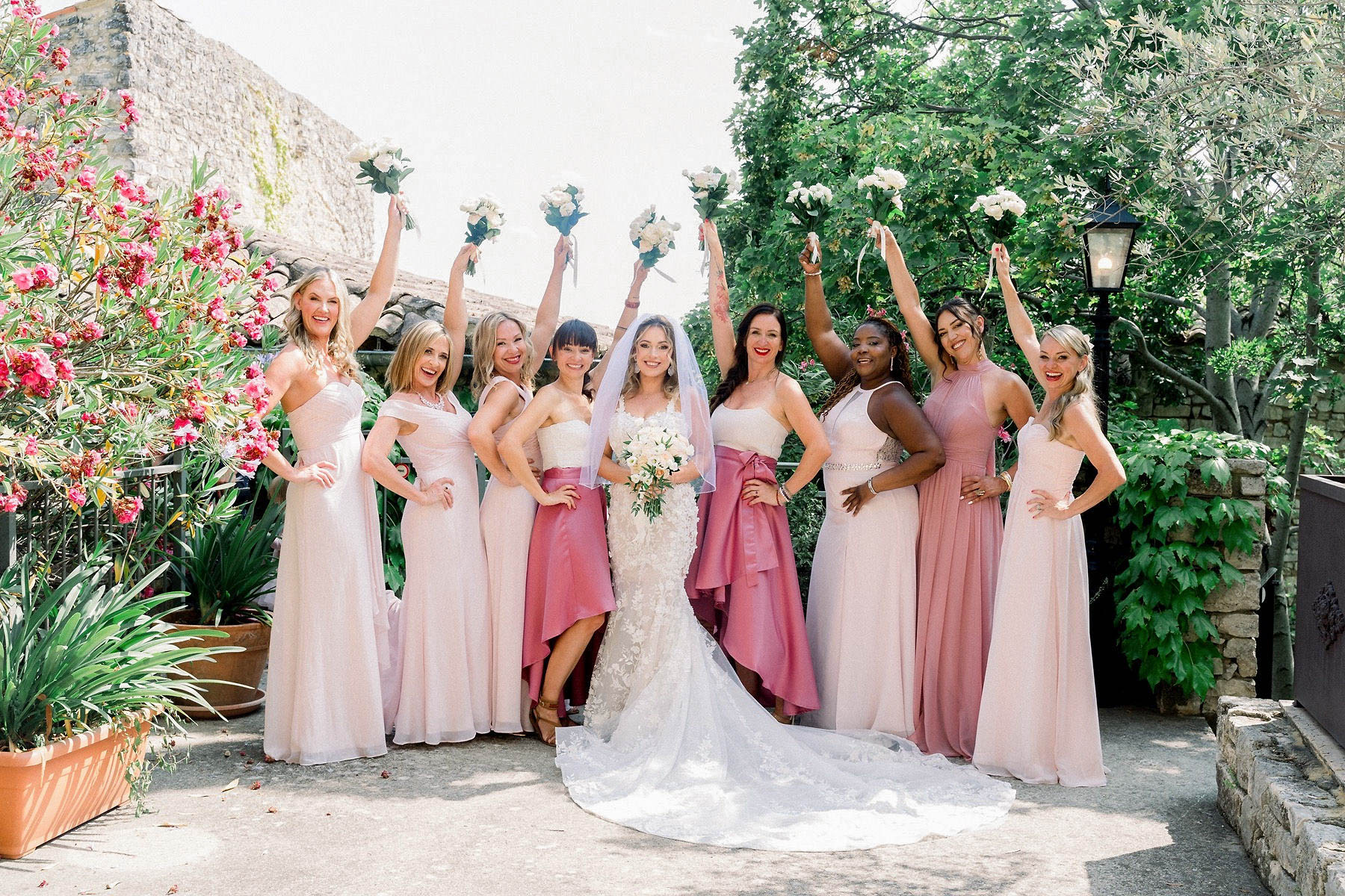 A bridal party portrait taken outdoors on a terrace or courtyard area of what appears to be a Provençal stone venue, with lush greenery and a lantern visible in the background. The bride stands at center in a fitted ivory lace gown with a cathedral-length train and a white veil, holding a bouquet of white and blush blooms. She is flanked by eight bridesmaids wearing a mix of blush pink and dusty rose floor-length gowns in varying styles — some in soft blush chiffon, two in deeper mauve-rose satin with high-low hemlines — all raising their white bouquets in the air in a celebratory pose. The color palette across the group is cohesive in the pink and rose family, creating a mismatched-but-coordinated look typical of a classic-romantic styling approach. Wide group shot taken at eye level in bright midday light.