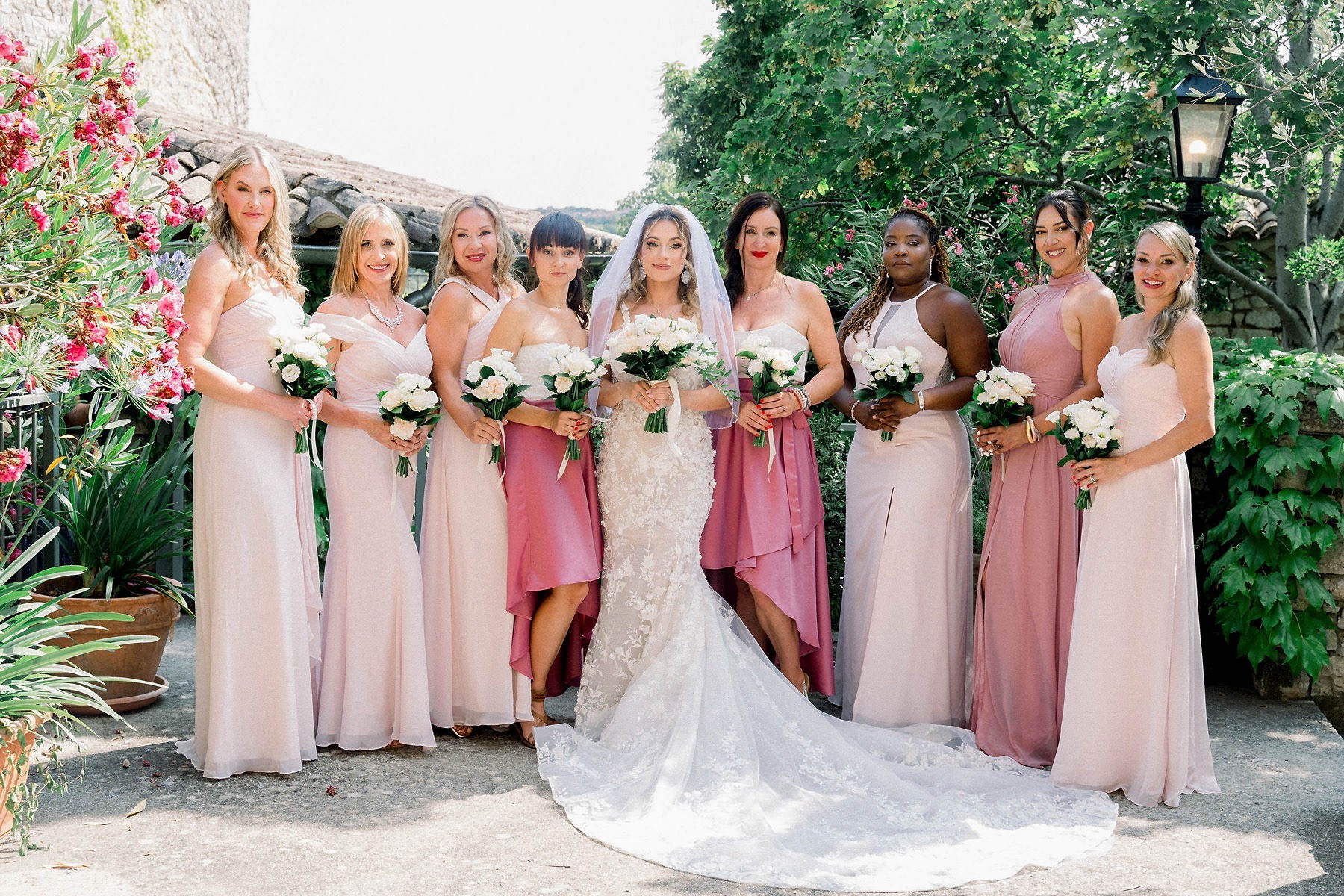 A bridal party portrait taken outdoors in a courtyard or terrace setting, likely at a French stone-building venue. The bride stands at center wearing a fitted floral lace gown with a cathedral-length lace-edged train and a cathedral veil, holding a bouquet of white roses and greenery. She is flanked by eight bridesmaids wearing a mix of blush pink floor-length gowns in various styles and two bridesmaids in deeper dusty rose/mauve high-low satin dresses, creating a tonal pink color palette across the group. All bridesmaids carry matching bouquets of white roses with dark green foliage. The composition is a wide group portrait shot in bright natural daylight, with the nine women lined up and facing the camera directly.