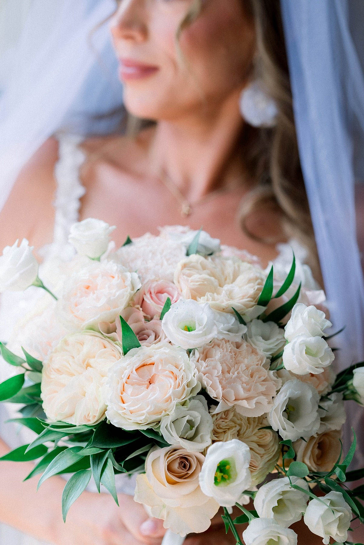 A close-up bridal portrait focused on the bouquet in the foreground, with the bride softly out of focus in the background. The rounded bridal bouquet features blush and cream garden roses, white lisianthus, soft pink spray roses, and carnations, accented with green ruscus and eucalyptus foliage. The bride is wearing a white lace-strap gown, a sheer veil, white drop earrings, and a delicate pendant necklace, with her blonde hair worn down in loose waves. The overall palette is blush, cream, and white with classic styling.