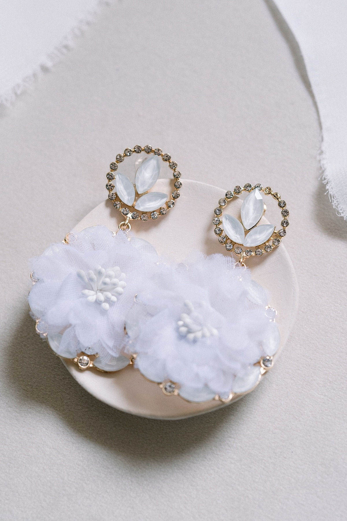 Close-up detail shot of a pair of bridal drop earrings displayed on a small cream ceramic dish against a soft grey background with a white silk ribbon partially visible. The earrings feature gold-tone circular stud tops set with small grey crystals and pale blue marquise-cut stones arranged in a leaf motif, from which hang large white fabric flower pendants made of layered organza or chiffon petals with white resin stamens at the center, edged with small gold-set crystal accents. The styling is classic bridal with a soft, white and gold palette. The composition is a styled flat-lay detail shot.