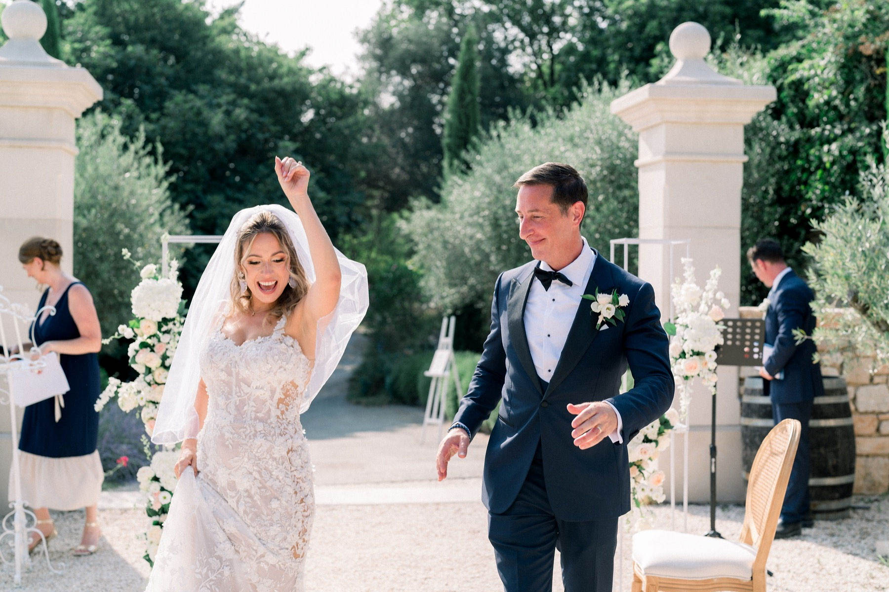 The bride and groom are captured mid-recessional immediately after their outdoor ceremony, walking between two white stone pillars that frame the ceremony exit. The bride, wearing a fitted lace gown with a cathedral veil, has one arm raised in celebration and is laughing expressively, while the groom in a navy tuxedo with black lapels and bow tie smiles beside her. Floral arrangements of white hydrangeas, white roses, and blush peach blooms on white metal stands flank the pillars, and the groom wears a matching white floral boutonniere. In the background, a woman in a navy midi dress and another guest in a navy suit are visible near a music stand, with a wooden barrel and gold Chiavari-style chairs also in frame. The setting is an outdoor garden courtyard with a classic, polished aesthetic, and the image is a candid wide shot full of genuine joyful energy.