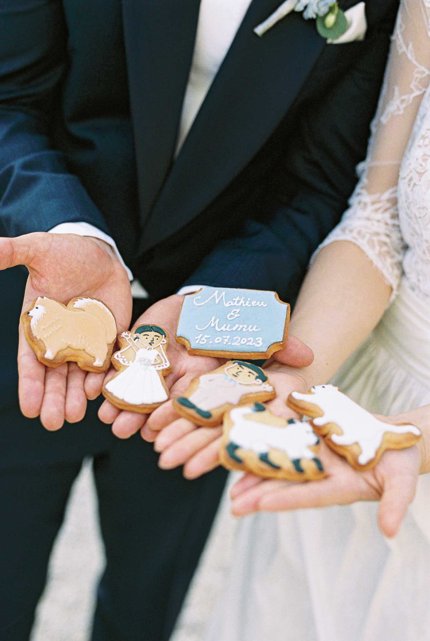 A close-up detail shot of a couple holding six custom royal icing decorated shortbread cookies in their outstretched hands. The cookies feature personalized designs including a bride figure in a white dress, a groom figure, a tan fluffy dog (likely a Samoyed or Chow Chow), a white polar bear or white dog, a tiger with teal and orange stripes, and a light blue plaque-shaped cookie reading 'Mathieu & Mumu 15.07.2023' in white script. The groom is wearing a dark navy suit with a green foliage boutonnière, and the bride is wearing a lace long-sleeve white gown with a tulle skirt. The cookies appear to be personalized wedding favors reflecting the couple's pets or meaningful symbols. The composition is a tight portrait-orientation detail shot focused on the hands and cookies.