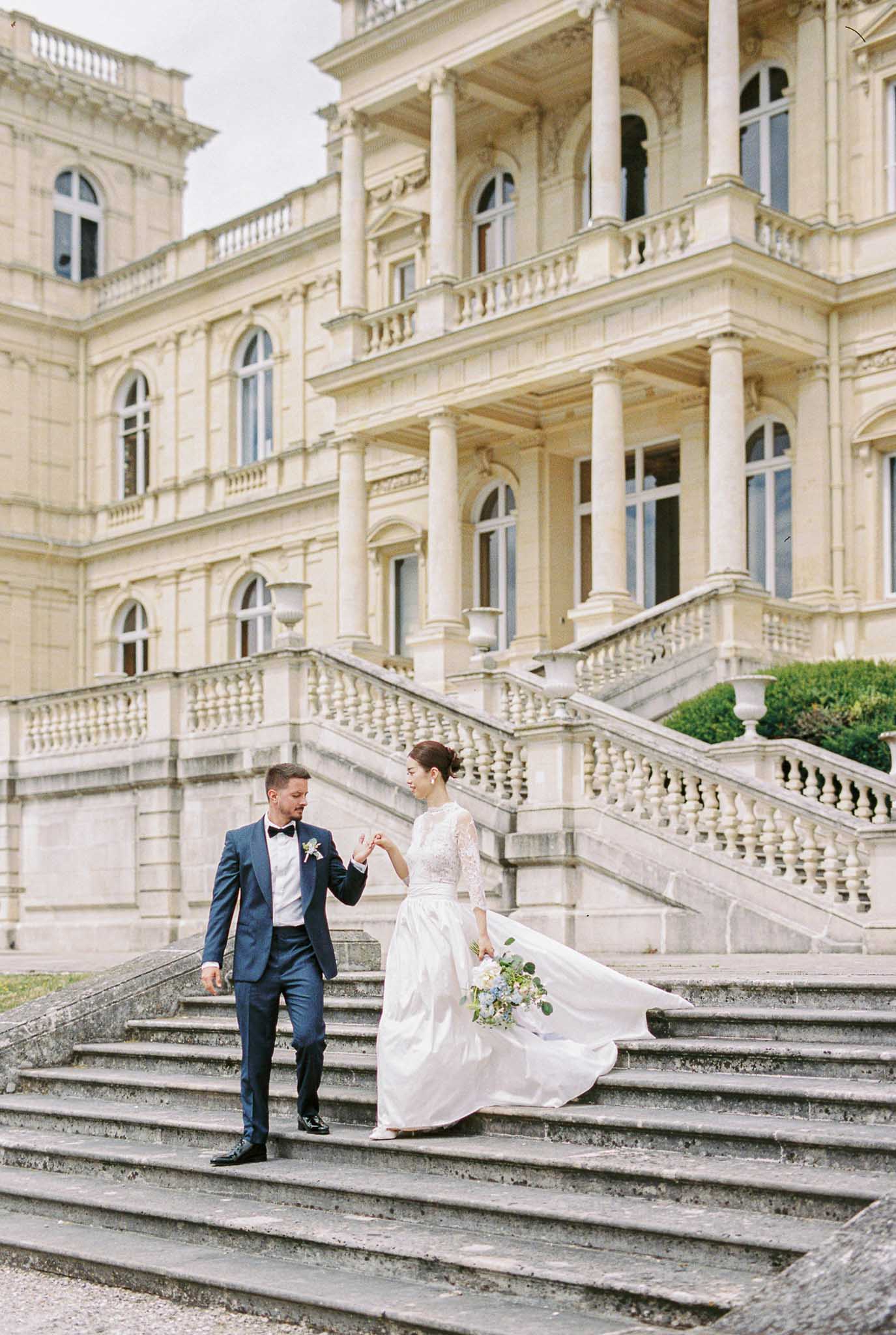 A couple portrait taken outdoors on the grand stone staircase of a large French château with classical architecture, featuring tall columns, arched windows, and ornate balustraded balconies across multiple stories. The groom, dressed in a navy blue tuxedo with a black bow tie and a small floral boutonnière, holds the bride's hand as they descend the steps together. The bride wears a two-piece white gown with a lace long-sleeve top and a full flowing skirt, with her hair styled in an updo, and she carries a loose bouquet of soft blue hydrangeas, white blooms, and eucalyptus. The shot is a medium-wide portrait capturing both figures in full length against the pale stone façade of the building. Potential venue feature image.