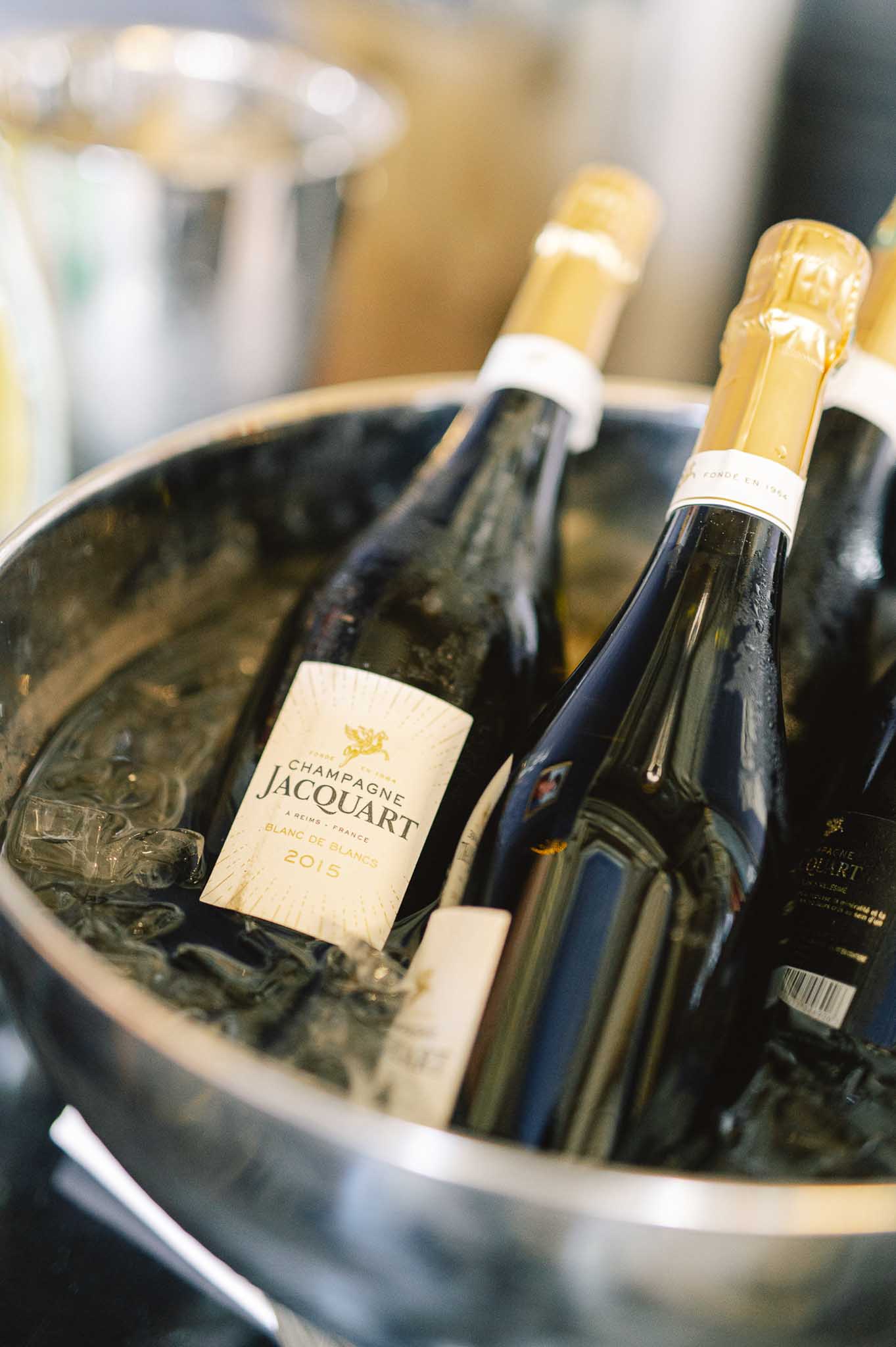 Close-up detail shot of multiple bottles of Champagne Jacquart Blanc de Blancs 2015 chilling in a stainless steel ice bucket filled with ice, likely during a cocktail hour or reception service. Three dark glass bottles with gold foil caps and cream-colored labels are visible, with a second ice bucket slightly out of focus in the background. The image is a tight, angled close-up with a shallow depth of field, emphasizing the champagne brand and the condensation on the bottles.