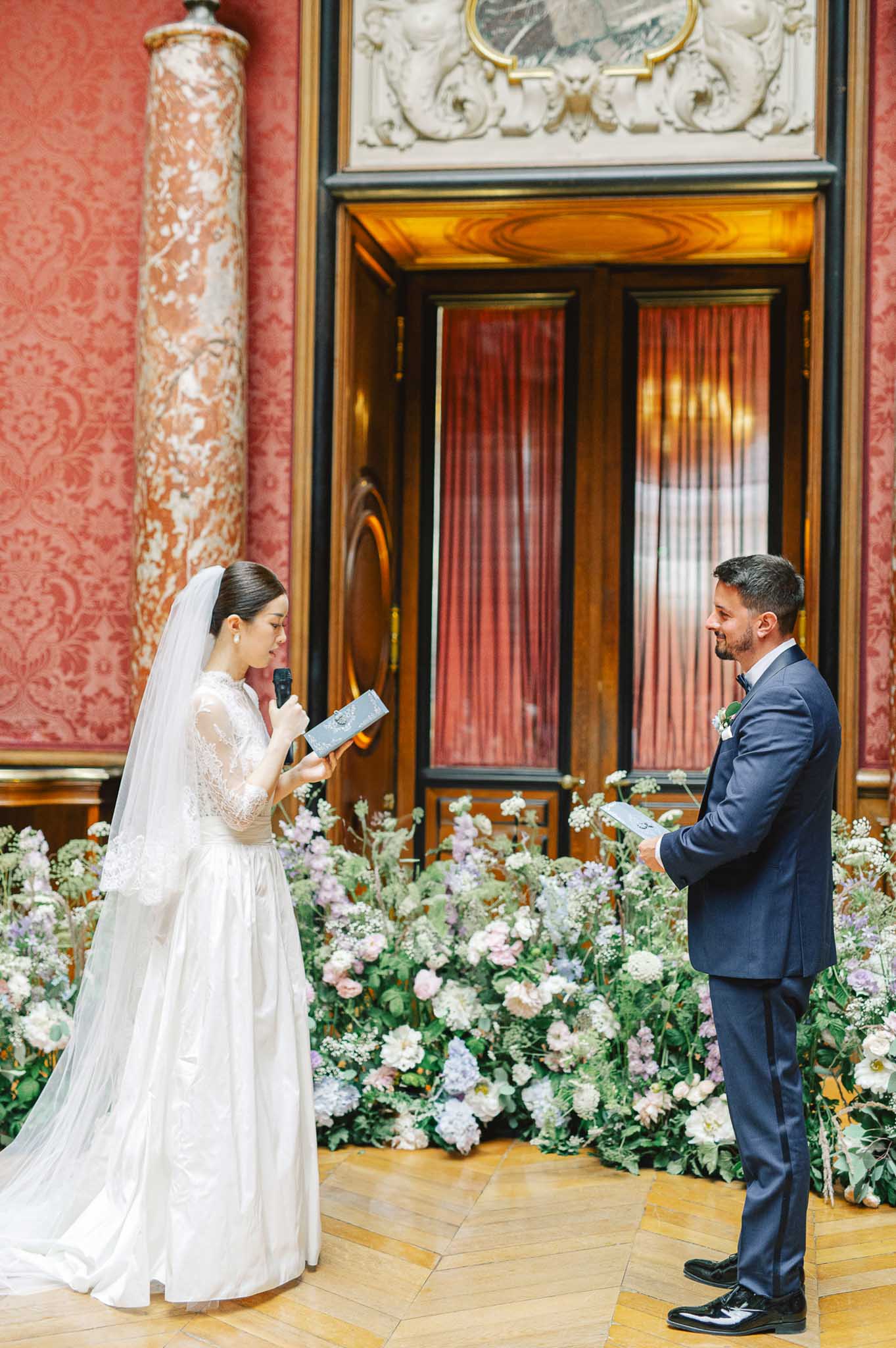 Bride reads vows to groom in Parisian hall with red damask walls and blush garden rose ground installation