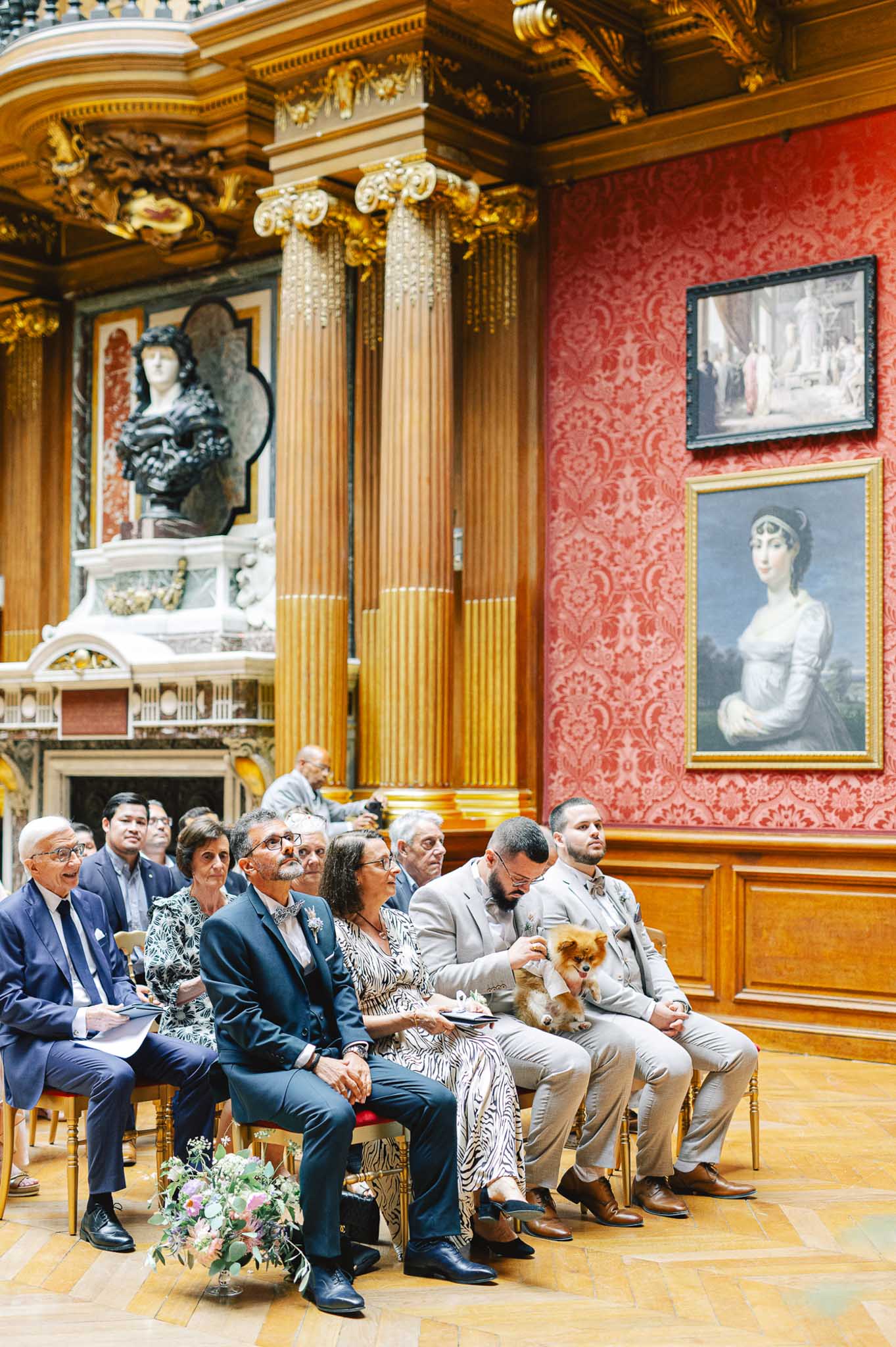 Wedding guests are seated during an indoor civil ceremony, attentively watching proceedings from gold-framed wooden chairs arranged in rows. The setting is a grand formal room featuring gold fluted columns with gilded Ionic capitals, deep red damask-patterned walls hung with large classical oil paintings, a marble fireplace with a dark stone bust, and a parquet wood floor — consistent with a French mairie or historic institutional salon. Approximately eight to ten guests are visible, dressed in smart-casual attire including a navy suit, a black-and-white patterned dress, and light beige suits; one guest is holding a small fluffy Pomeranian dog. A loose bouquet of mixed soft pink, lavender, and white blooms with greenery rests on the floor in the foreground. The shot is a wide-angle perspective taken from a low angle looking along the row of seated guests toward the front of the room. Potential venue feature image.