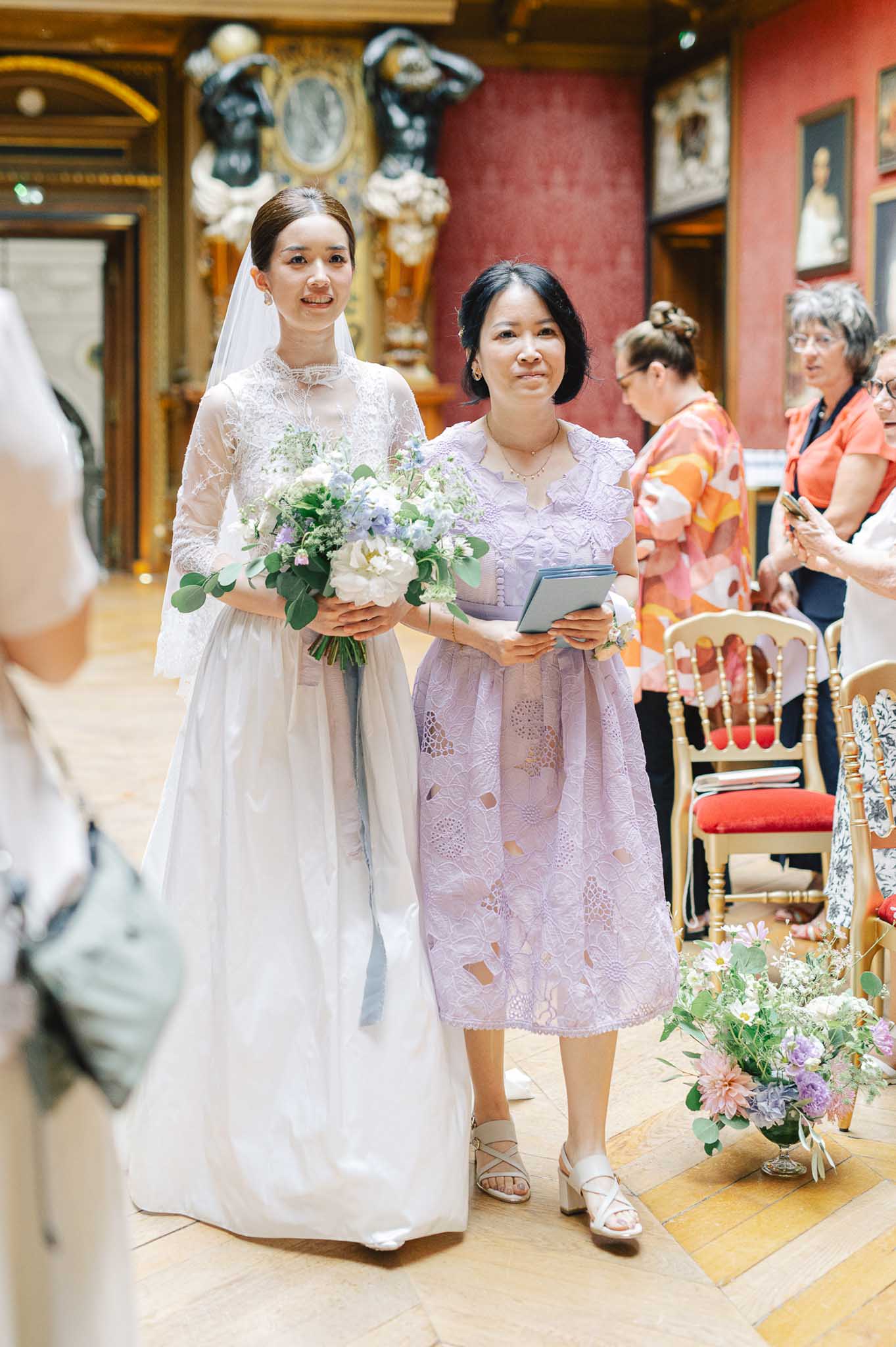 The bride is walking down the aisle during an indoor ceremony, accompanied by a woman in a lavender lace midi dress who appears to be a family member. The ceremony takes place inside a grand historic venue featuring deep red walls hung with oil portraits, ornate gold architectural details, dark bronze sculptures, and a parquet floor — consistent with a French château or palace interior. Gold Chiavari chairs with red cushions line the aisle, and low floral arrangements of pink dahlias, lavender blooms, white daisies, and eucalyptus mark the aisle edge. The bride wears a white gown with a high neckline, sheer lace long sleeves, and a full skirt, paired with a cathedral-length veil and her hair pulled back sleekly. She carries a loose, garden-style bouquet of white peonies, pale blue delphiniums, small white filler flowers, and eucalyptus. Several guests stand in the background, and a mid-shot composition captures both women walking toward the camera.