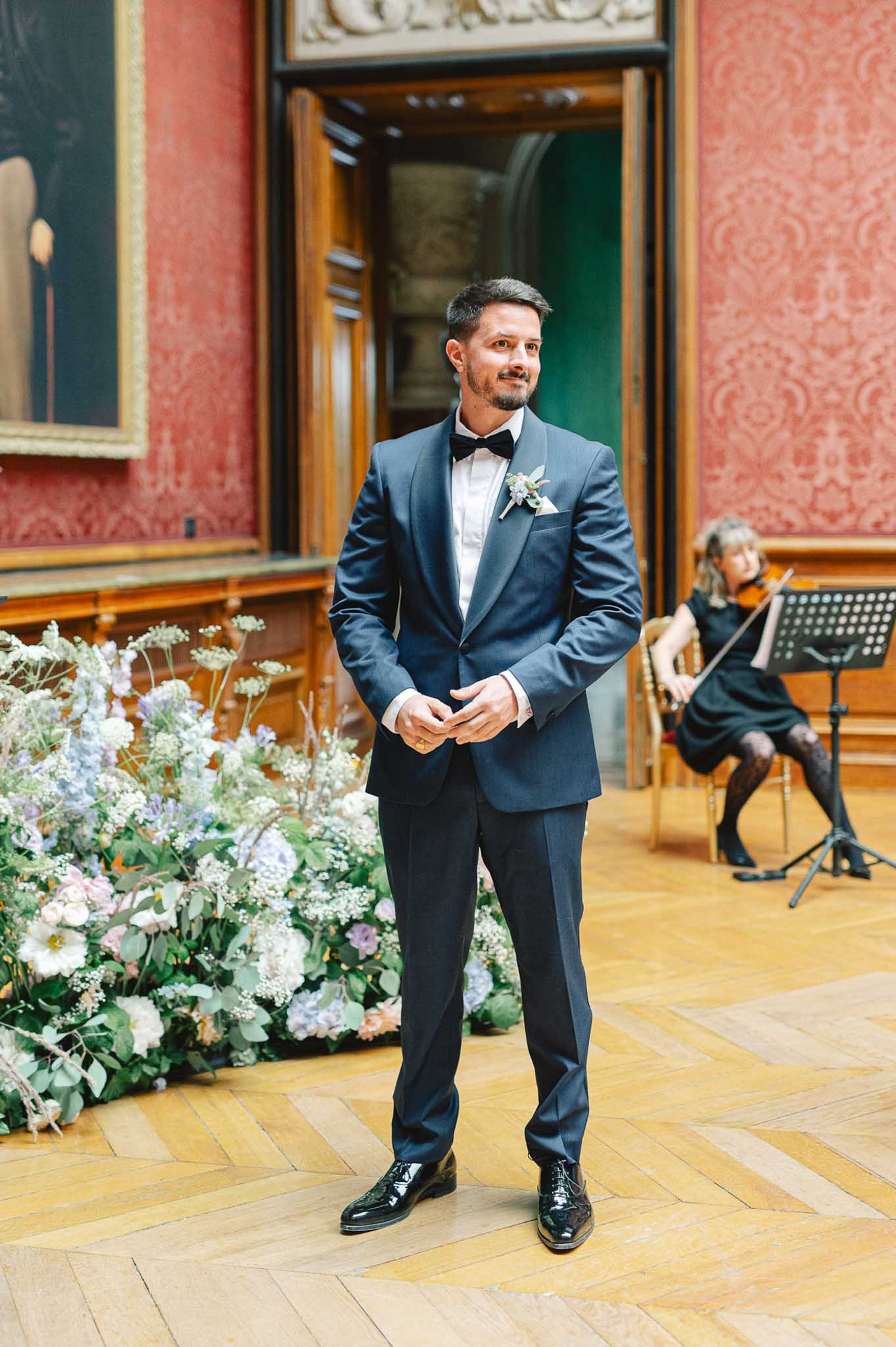 Groom in navy tuxedo at altar before white blush and lavender floral installation with violinist