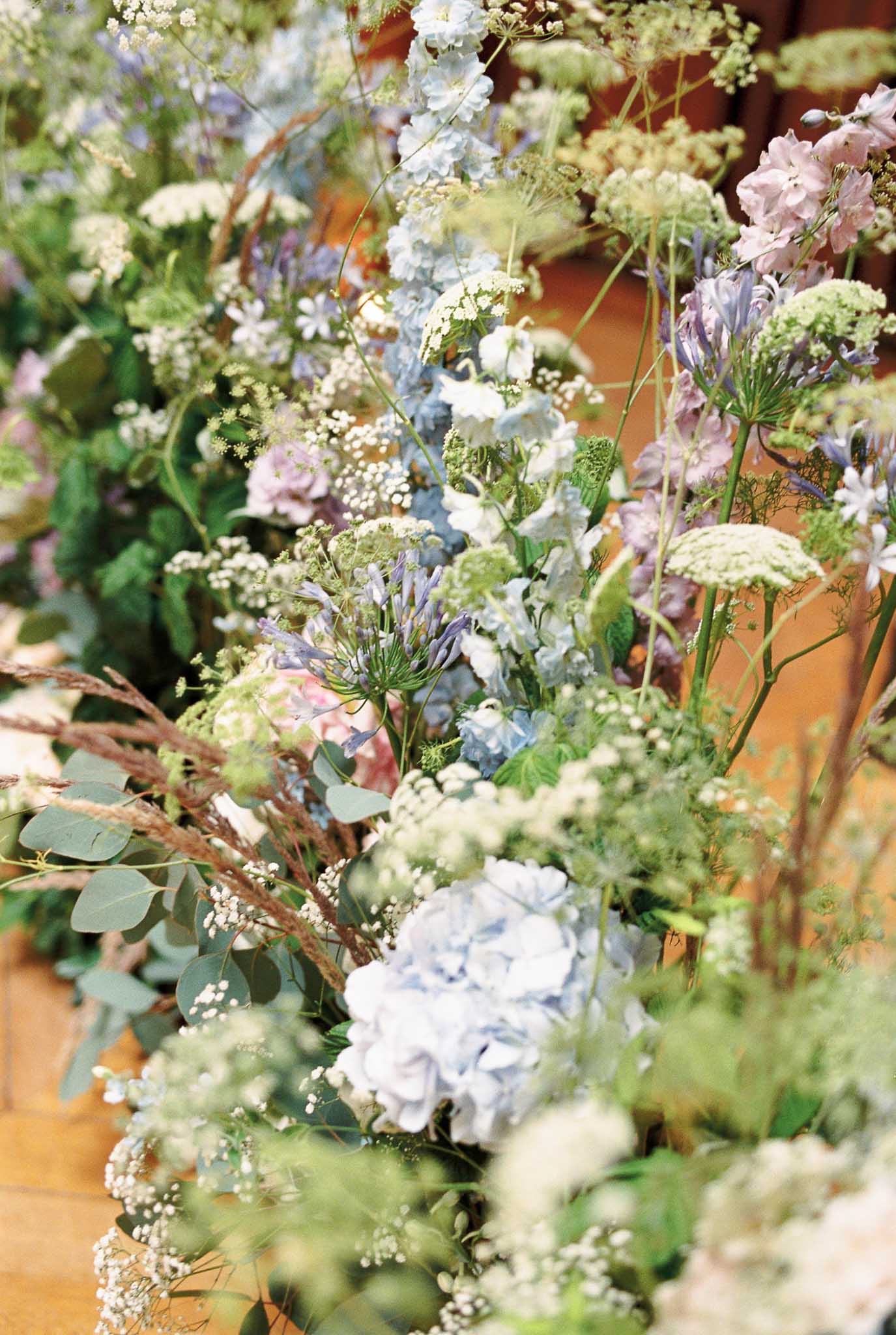 A close-up detail shot of a large-scale wedding floral installation arranged along what appears to be a wooden floor or low surface indoors, likely at a ceremony or reception venue. The arrangement features a soft, garden-style palette of pale blue hydrangeas, blue and mauve delphiniums, purple agapanthus, white queen anne's lace, gypsophila, and soft pink blooms, interspersed with eucalyptus, dried ornamental grasses, and fine green foliage. The overall styling is loose and wildflower-inspired, with tall stems extending upward to create an unstructured, meadow-like effect. The composition is a tight angled shot taken at floor level, with shallow depth of field that softly blurs the background.