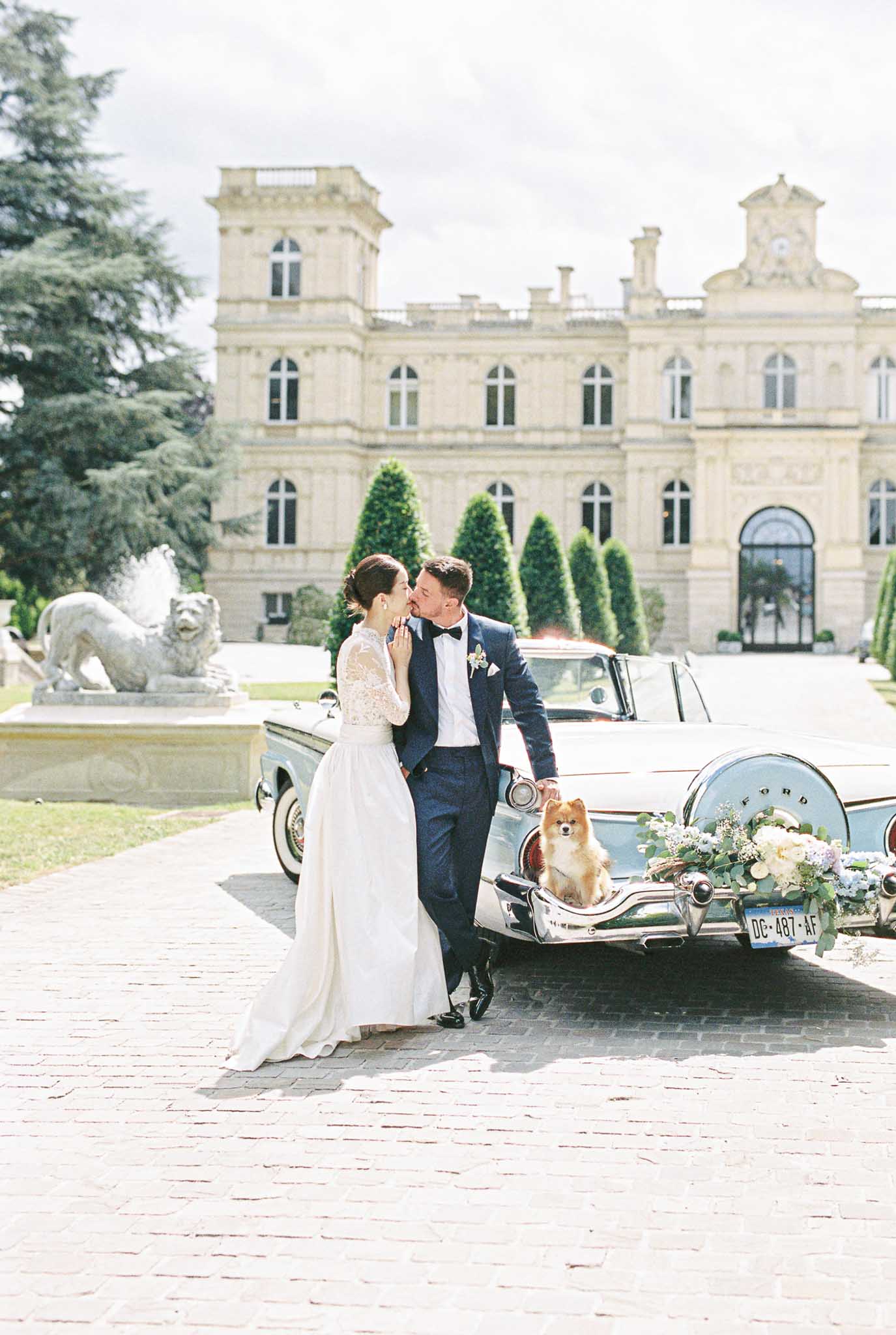 Bride and groom leaning against vintage blue Ford convertible on chateau forecourt with topiary and stone facade