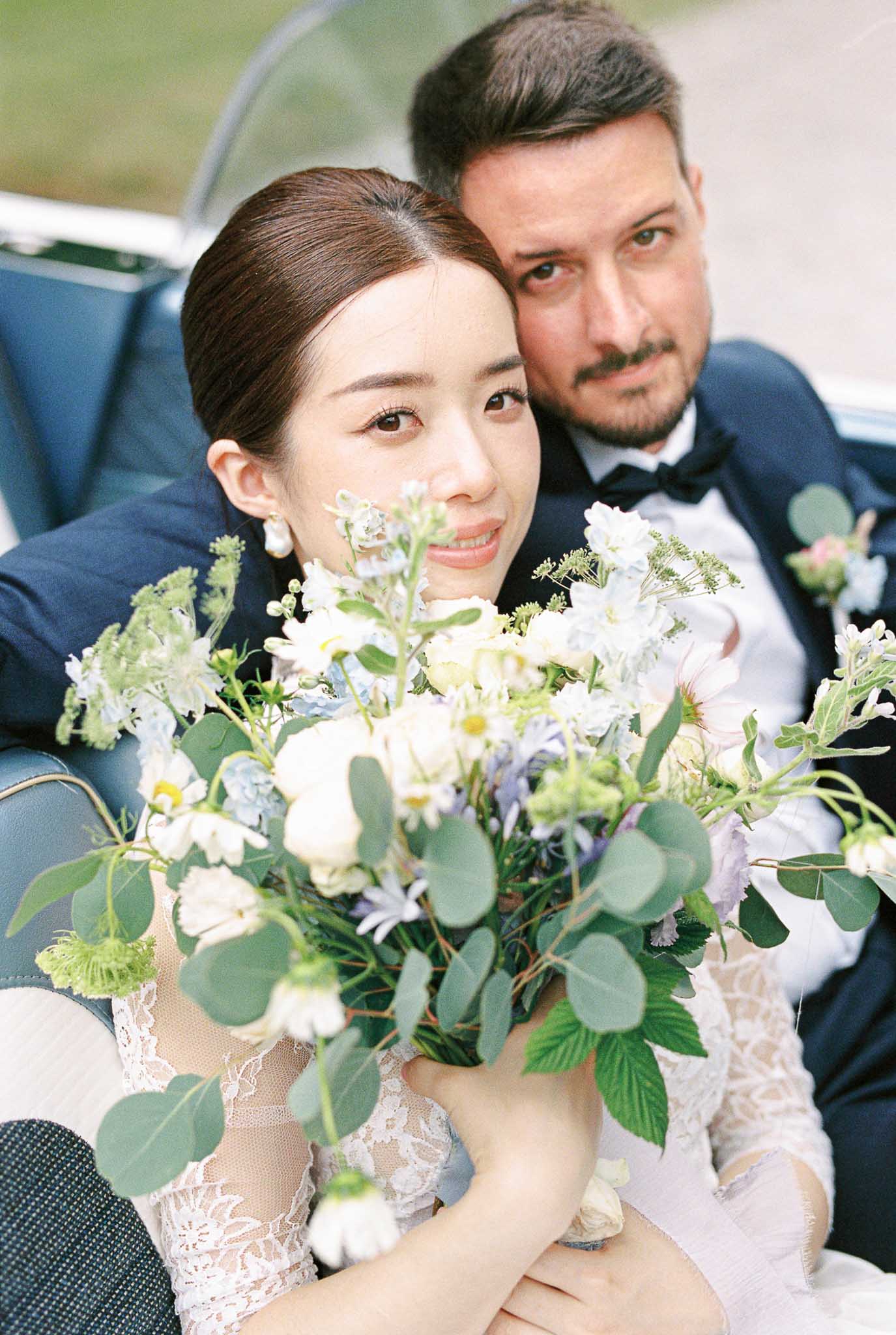 Bride and groom seated in vintage convertible with teal upholstery, bride holding white and pale blue bouquet