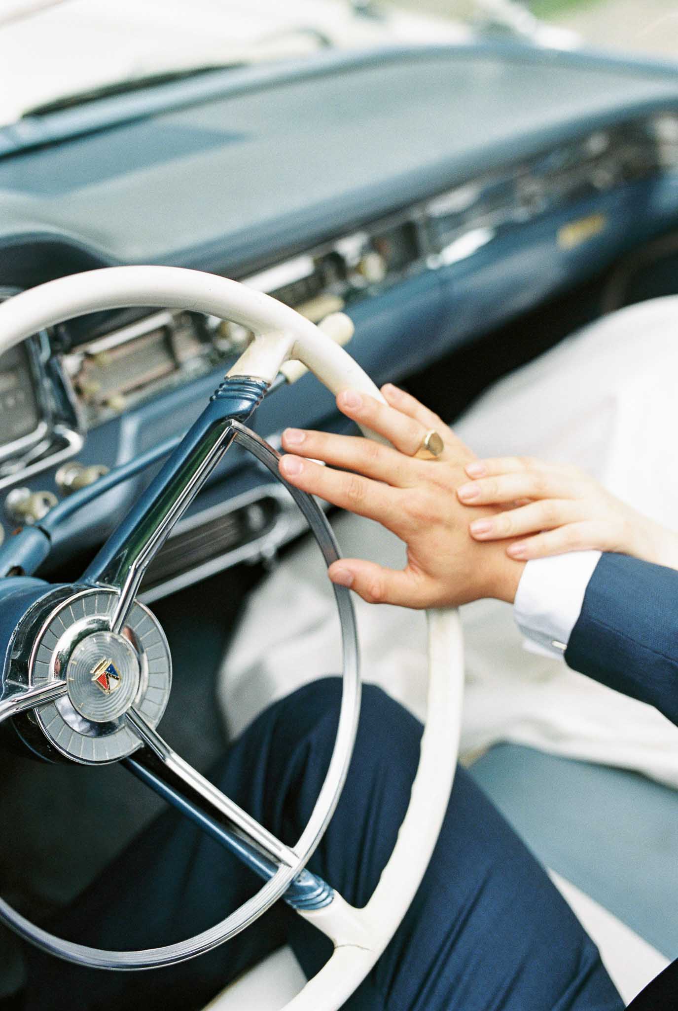 Two hands resting on vintage 1950s car steering wheel with gold signet ring and navy suit cuffs