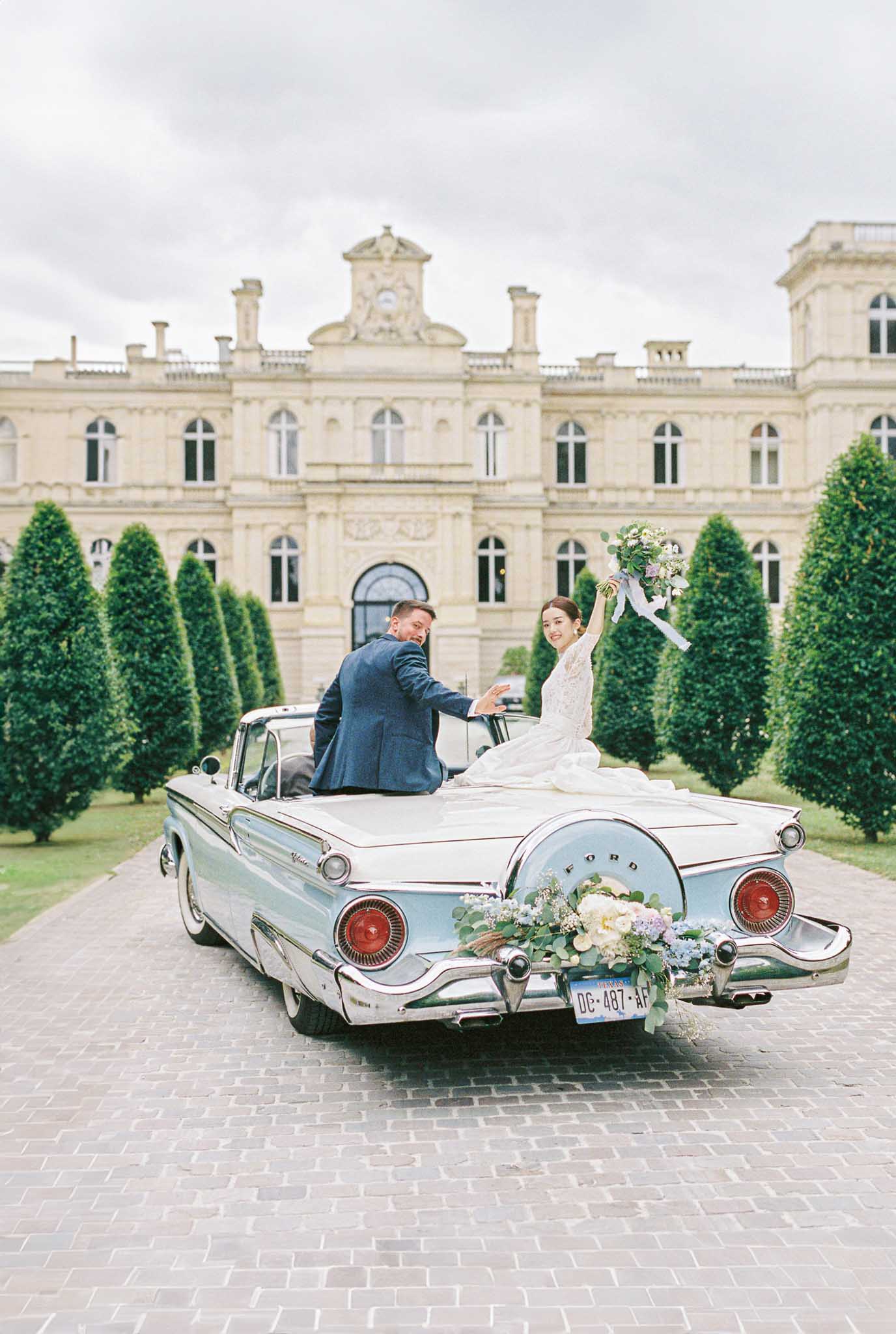 Bride and groom in vintage blue Ford convertible in front of chateau, bride raising bouquet