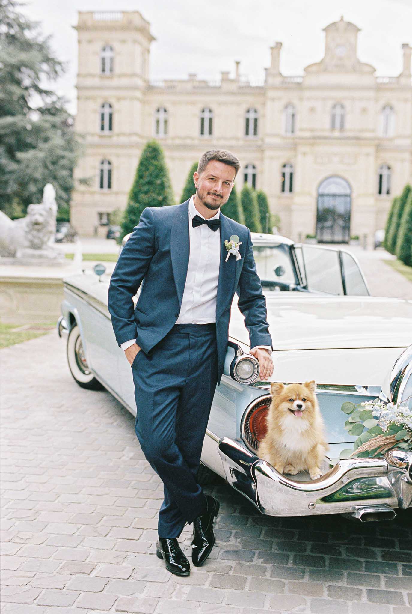 A groom portrait taken outdoors on a cobblestone driveway in front of a large French château with classical stone architecture. The groom is wearing a navy blue tuxedo with a black bow tie, white dress shirt, patent leather black shoes, and a boutonniere featuring small white flowers and eucalyptus. He is leaning casually against a vintage pale blue American car with chrome detailing, which is decorated with a floral arrangement of eucalyptus and light blue blooms on the rear bumper. A small golden Pomeranian dog sits on the car's rear bumper beside him. The composition is a full-length portrait shot with the château facade and manicured topiary trees visible in the background. Potential venue feature image.