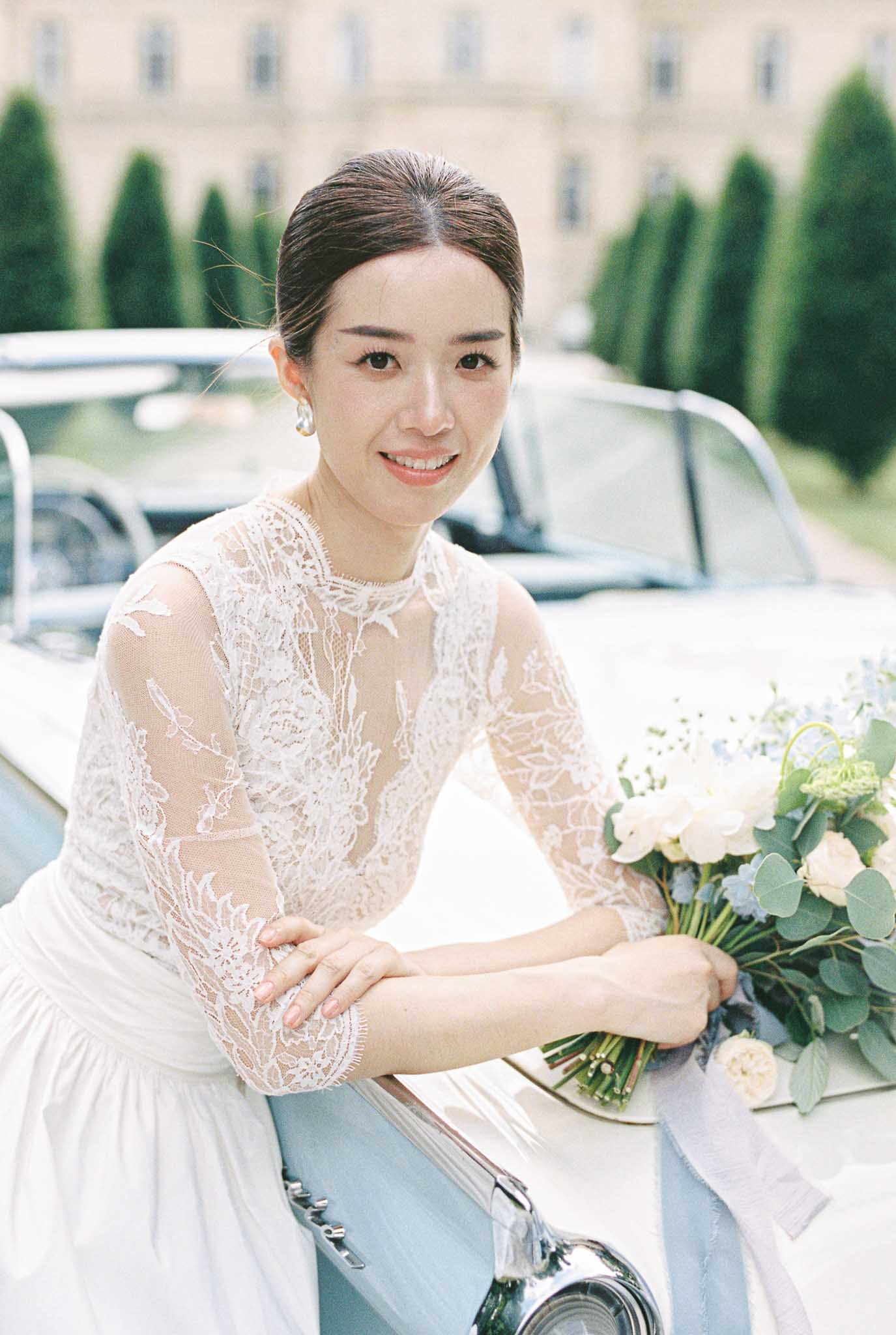 Bride leaning against pale blue vintage convertible holding bouquet of white roses, orchids, and blue flowers