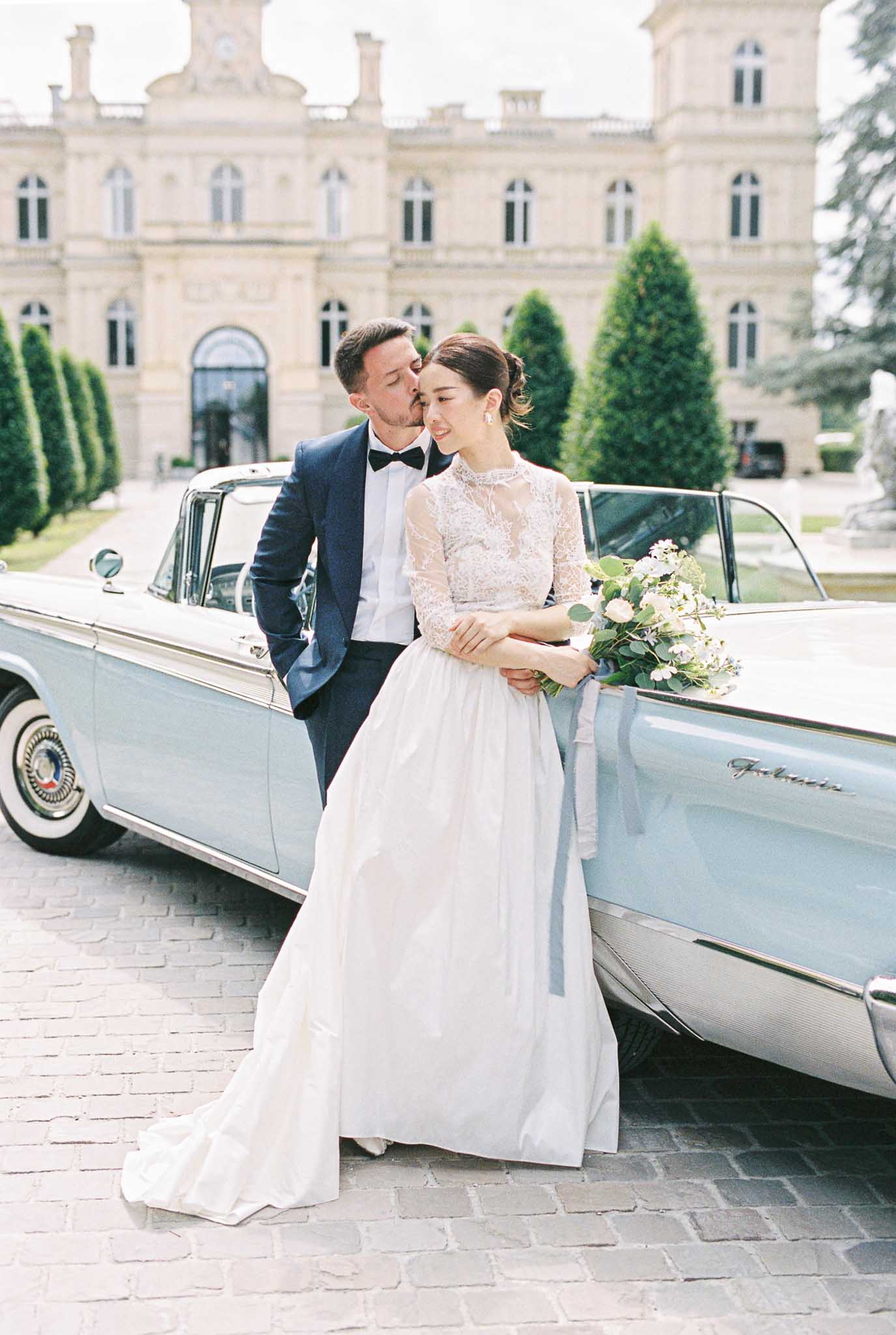 Groom kissing bride's cheek beside powder-blue vintage Ford convertible on chateau cobblestone forecourt