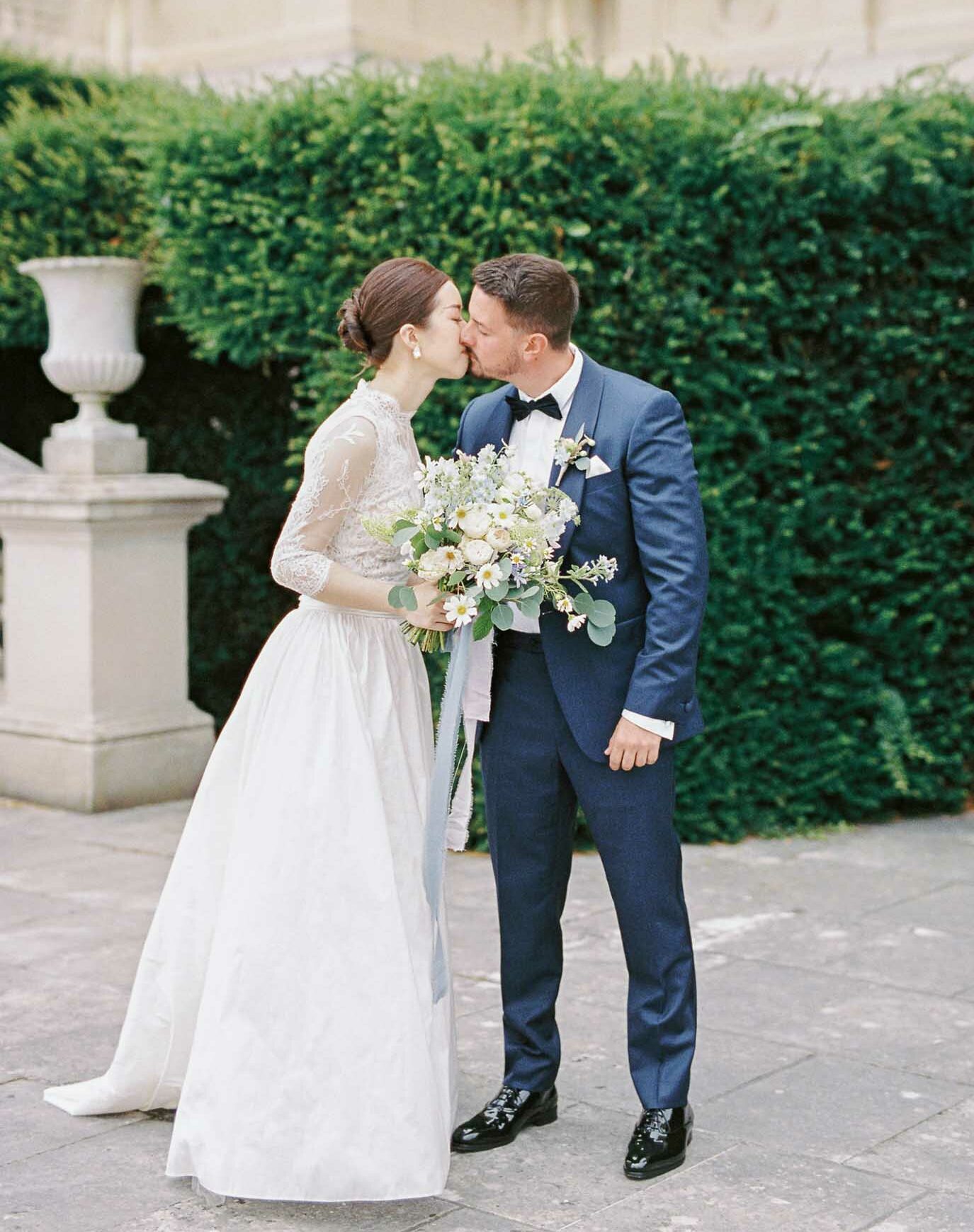 A couple portrait taken outdoors on a stone terrace, with the bride and groom sharing a kiss. The bride wears a two-piece ivory gown with a lace three-quarter-sleeve top featuring a high neckline and an a-line skirt, her dark hair pinned up in a low bun with pearl drop earrings. She holds a loose, garden-style bouquet of white ranunculus, white daisies, dusty blue and lavender blooms, and eucalyptus with trailing pale blue silk ribbon. The groom wears a navy blue suit with a black bow tie and a small boutonniere of greenery and a white bloom. A clipped formal hedge runs across the background, with a stone decorative urn visible to the left, suggesting a formal French château or estate garden setting. The shot is a medium full-length portrait with a classic, clean aesthetic.