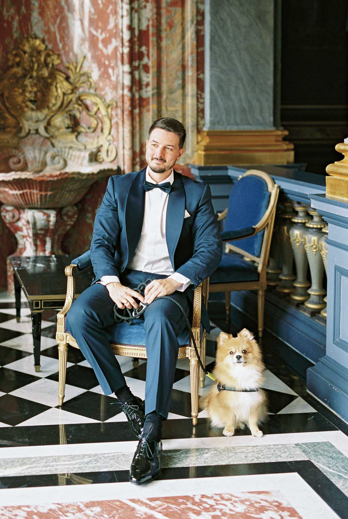 Groom in navy tuxedo seated in gold Louis XVI chair with Pomeranian dog on checkerboard marble floor