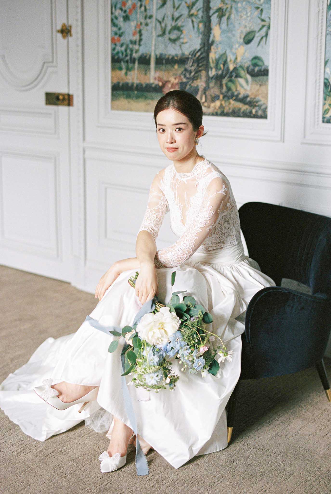 A bridal portrait taken indoors in a classically decorated room featuring white wood-paneled walls and a large decorative tapestry painting in the background. The bride is seated on the floor leaning against a navy blue velvet armchair, wearing a white gown with a sheer lace long-sleeve bodice and a voluminous silk skirt with a short train, paired with white heeled shoes adorned with lace or floral detailing. She holds a loosely arranged bouquet of ivory peonies, pale blue delphinium, white wildflowers, and eucalyptus tied with a dusty blue silk ribbon. Her dark hair is pulled back into a low bun, and she wears small drop earrings. The image is shot in a medium portrait format with soft, natural window light and a classic, polished interior styling.