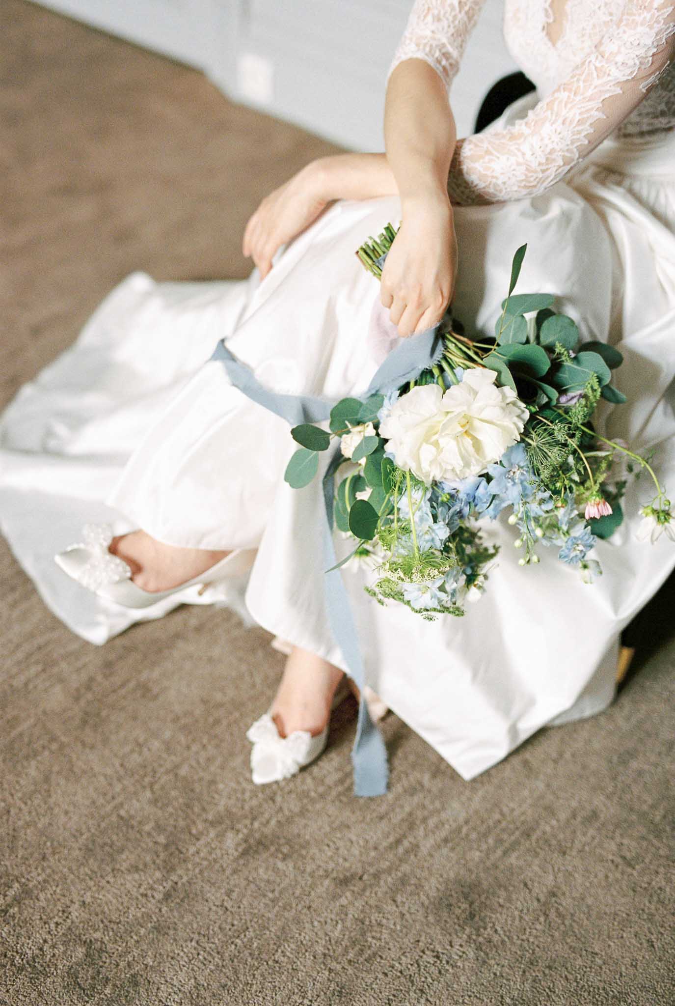 A getting-ready or bridal portrait detail shot of a seated bride, framed from the torso down. She is wearing a white satin skirt with a lace long-sleeve bodice and white embellished pointed-toe flats with a floral bow detail. She holds a loose, garden-style bridal bouquet featuring ivory garden roses, soft blue delphinium, eucalyptus, Queen Anne's lace, and small pink accent blooms, tied with a long dusty blue silk ribbon. The composition is a close-up portrait taken from above, shot on film with a soft, slightly warm tone and shallow depth of field.
