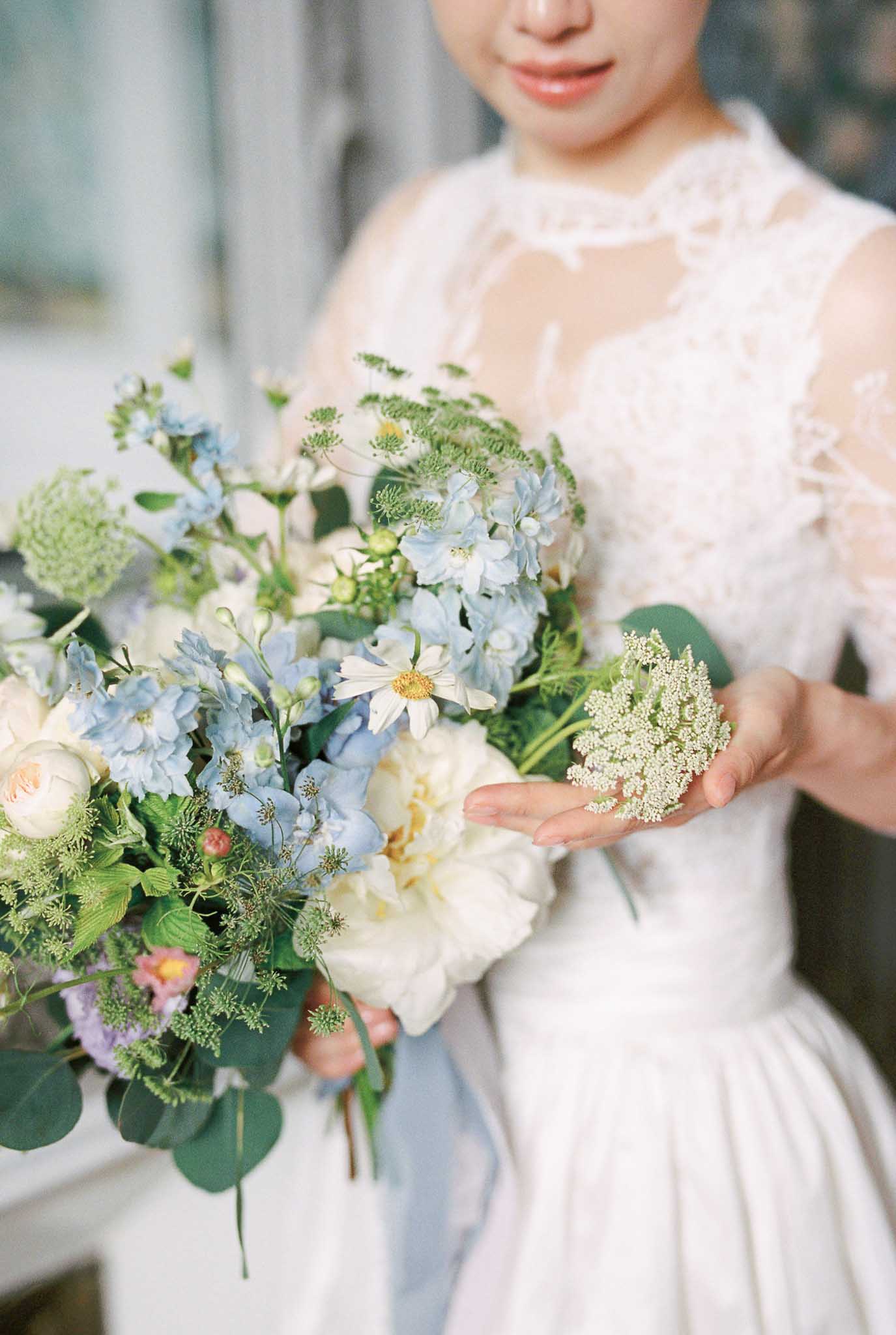 Bride in embroidered lace gown holding garden bouquet of blue delphiniums, cream peonies, and cosmos