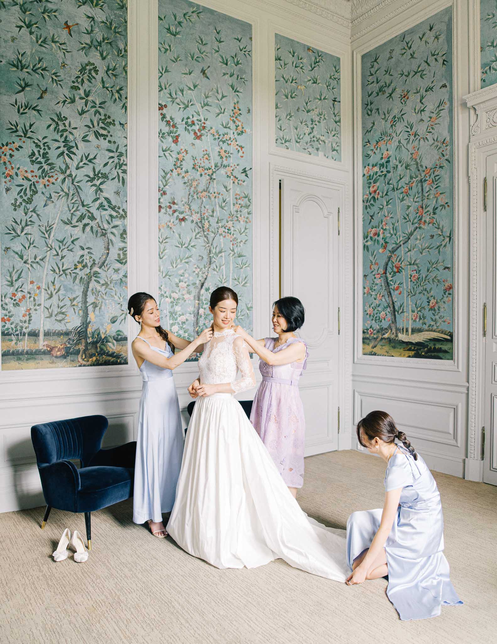 Three attendants in blue and lavender dresses helping bride into lace gown in chinoiserie room
