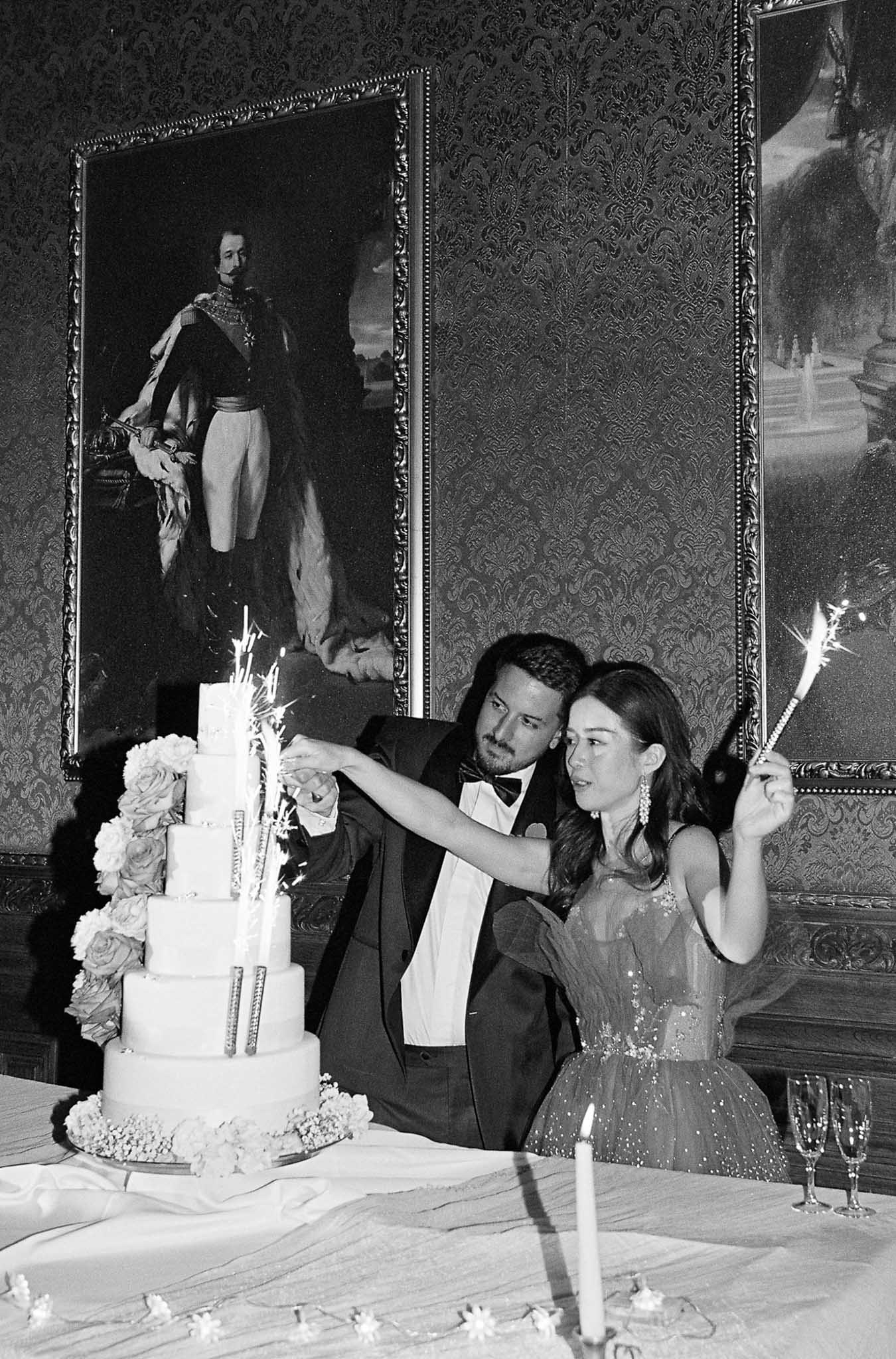 Bride and groom cutting four-tier cake with sparkler fountains in chateau ballroom with gilt paintings in B&W
