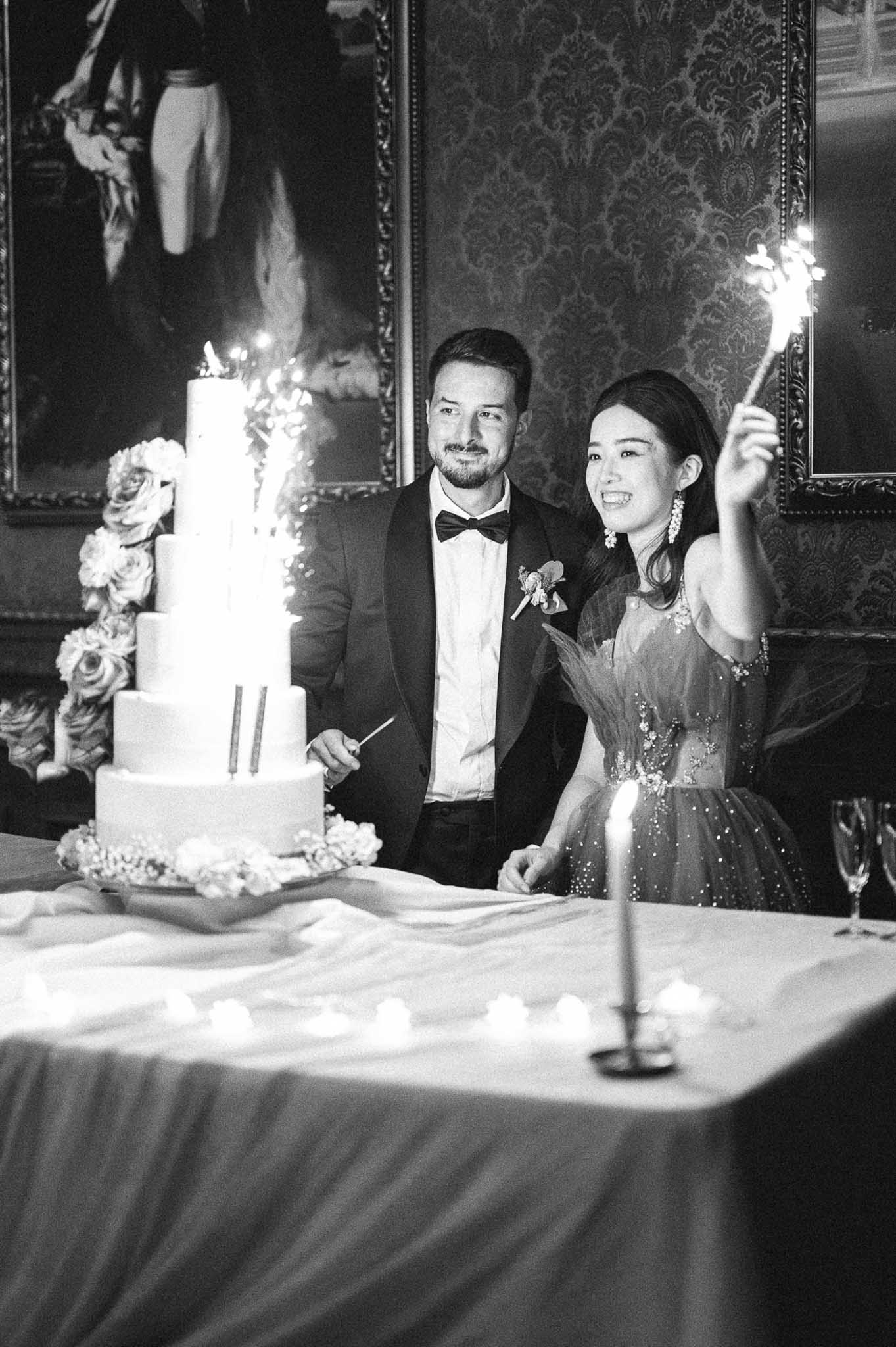 Black-and-white shot of couple behind five-tier wedding cake with sparklers in ornate chateau interior