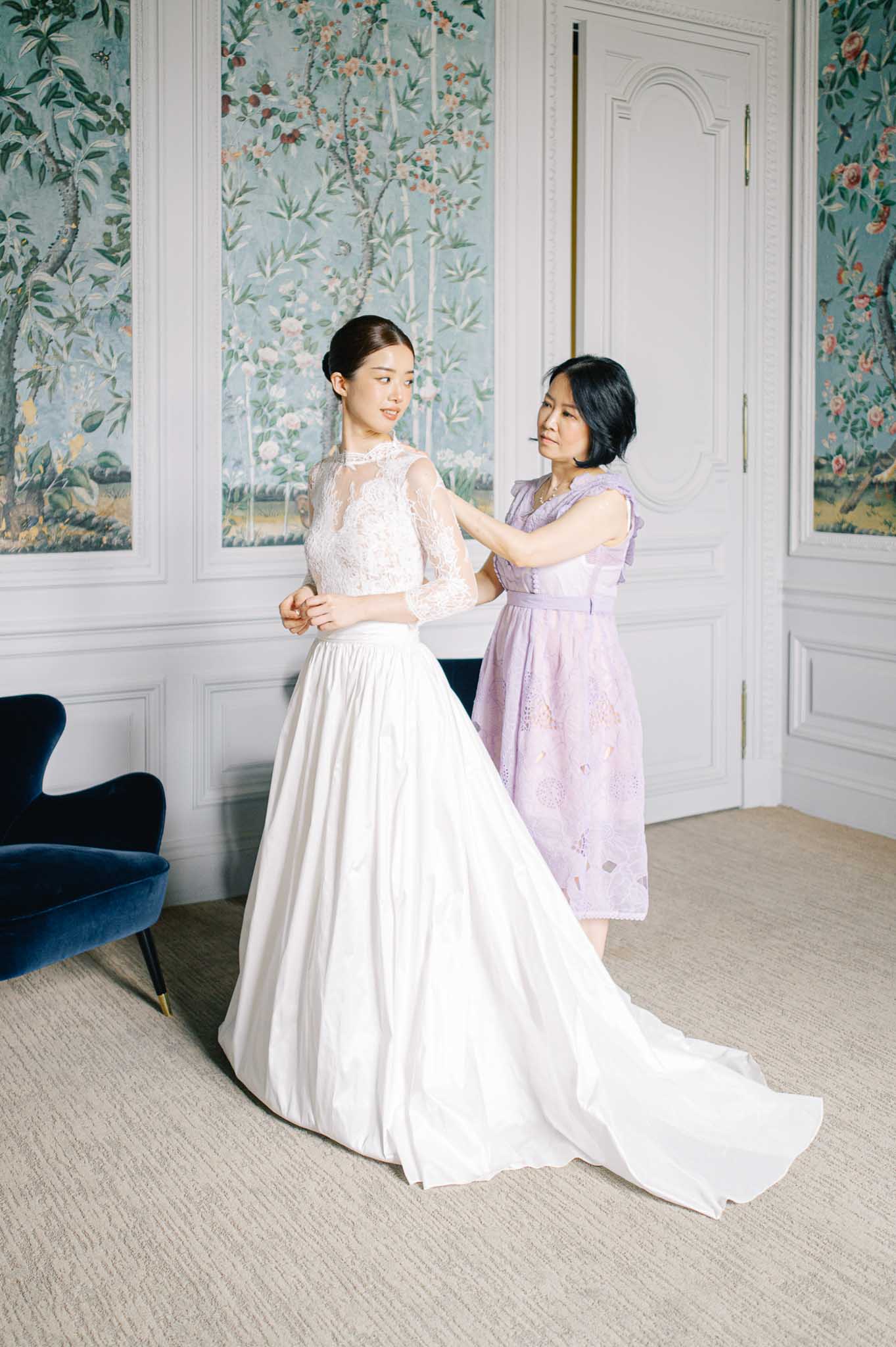 Mother helping bride into two-piece lace illusion top and satin skirt in chinoiserie-panelled chateau room