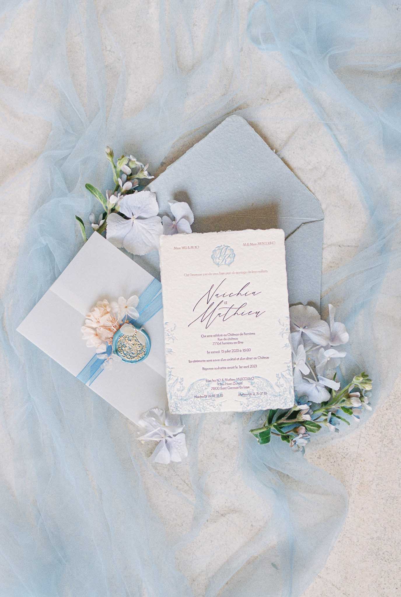 French wedding invitation suite with navy calligraphy on cream card, blue wax seal, and hydrangea petals on tulle