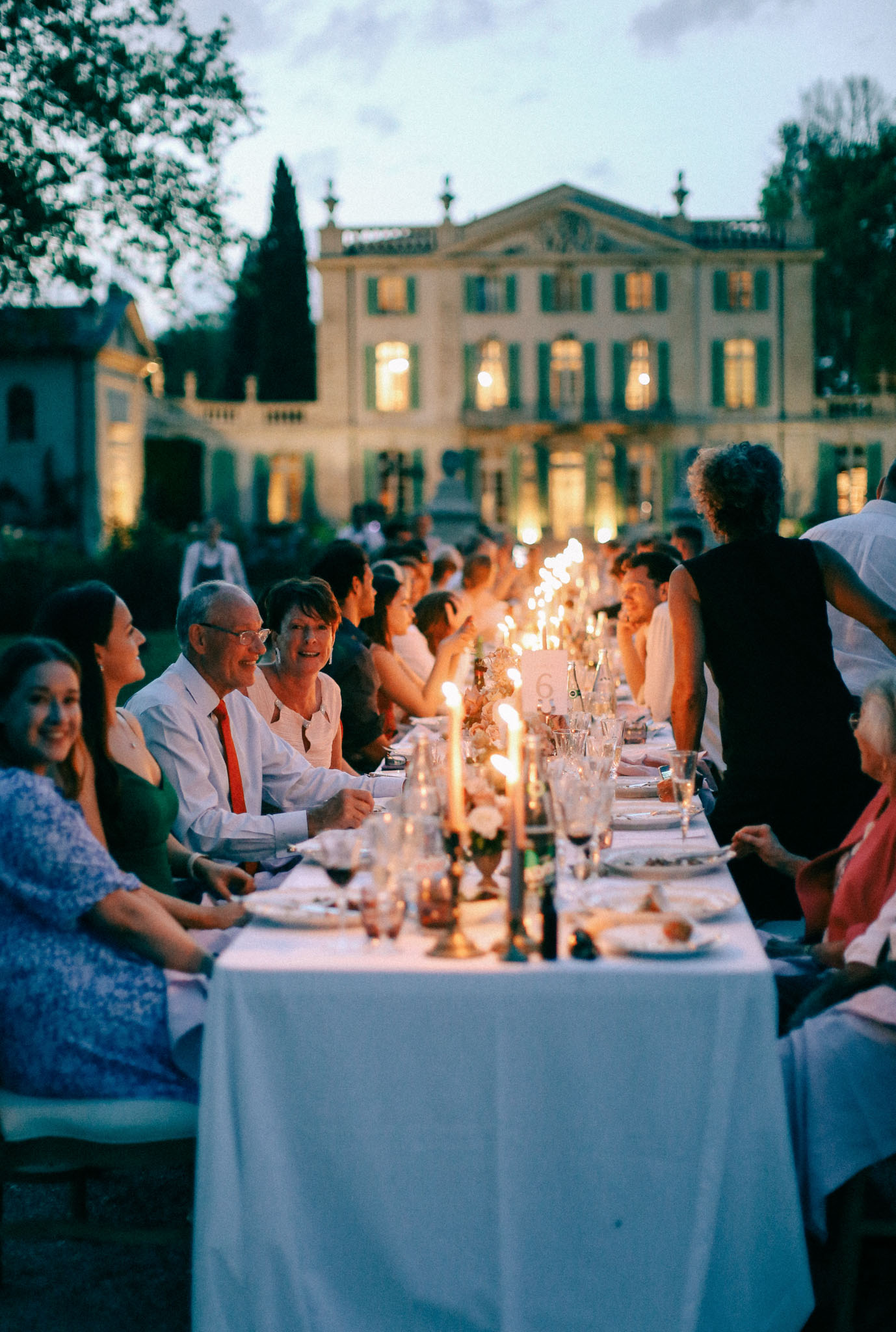 An outdoor wedding reception dinner is underway at dusk, with a long banquet table seating approximately 20–30 guests on both sides. The table is dressed with a white linen cloth and styled with tall taper candles, small floral arrangements featuring white blooms, crystal glassware, and white dinner plates. Guests are dressed in a mix of smart casual attire — notably a man in a white shirt with a red tie, a woman in a green dress, and another in a blue floral dress in the foreground. The warm candlelight contrasts with the cool blue dusk sky, creating a moody evening atmosphere. In the background stands a large French manor house or château with teal-green shutters, classical architectural detailing, and warm interior lighting glowing from its windows, flanked by uplighting at its base. Wide shot taken from a low angle along the length of the table, with the château centered in the soft-focus background. Potential venue feature image.