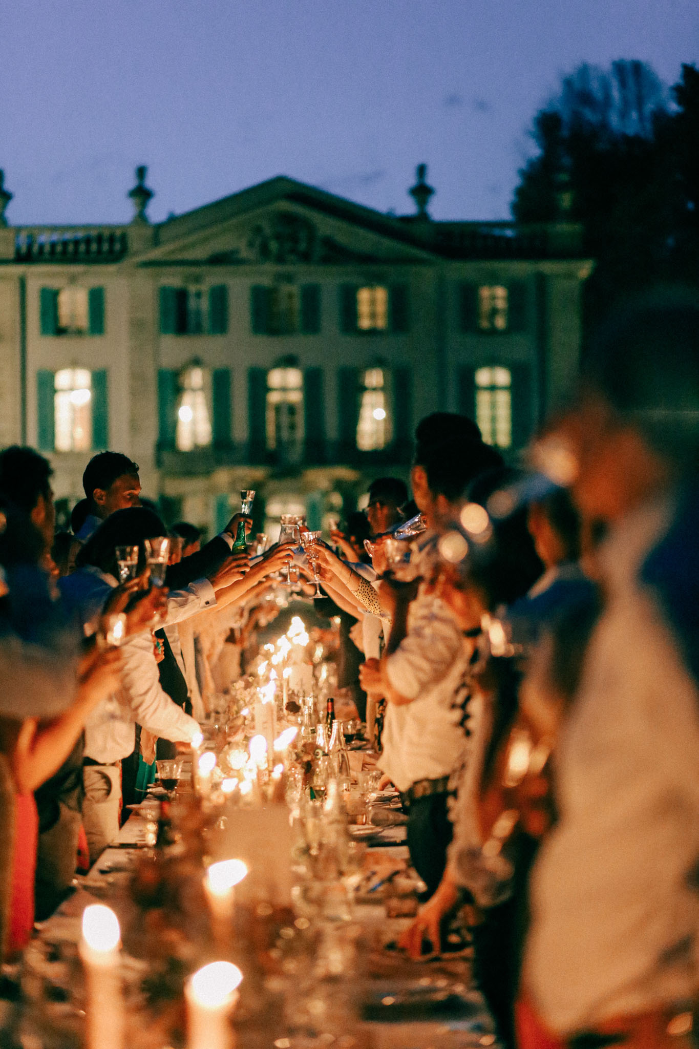 An outdoor evening reception toast is taking place at a long candlelit dining table, with approximately 20–30 guests raising their glasses simultaneously. The table is lined with numerous candles and glassware, casting warm golden light along its length. The setting is the grounds of a large classical French manor or château with green shutters and lit interior windows visible in the background against a deep blue dusk sky. Guests are dressed in smart-casual attire, with several in white shirts. The shot is taken from a low angle along the table, with foreground candles and a blurred guest creating a shallow depth-of-field effect, placing the toast moment at the center of focus.